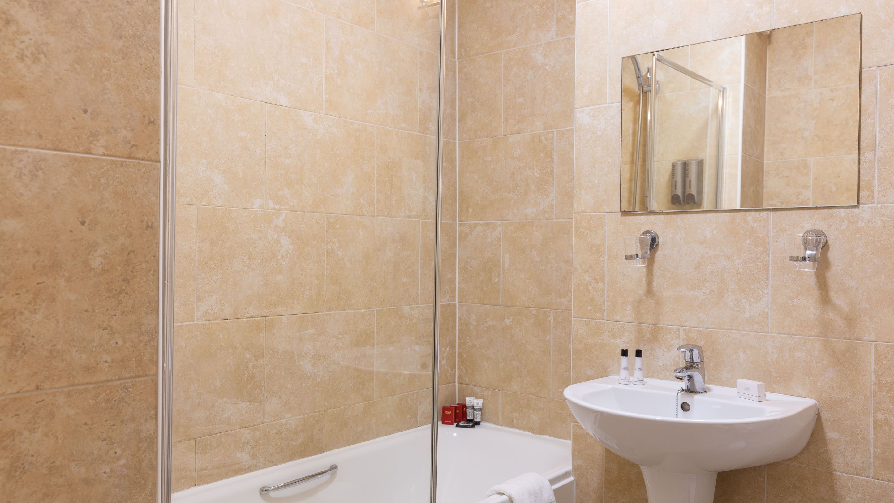 Un baño con baldosas color beige, cabina de ducha de vidrio, bañera blanca y lavamanos blanco con espejo superior.