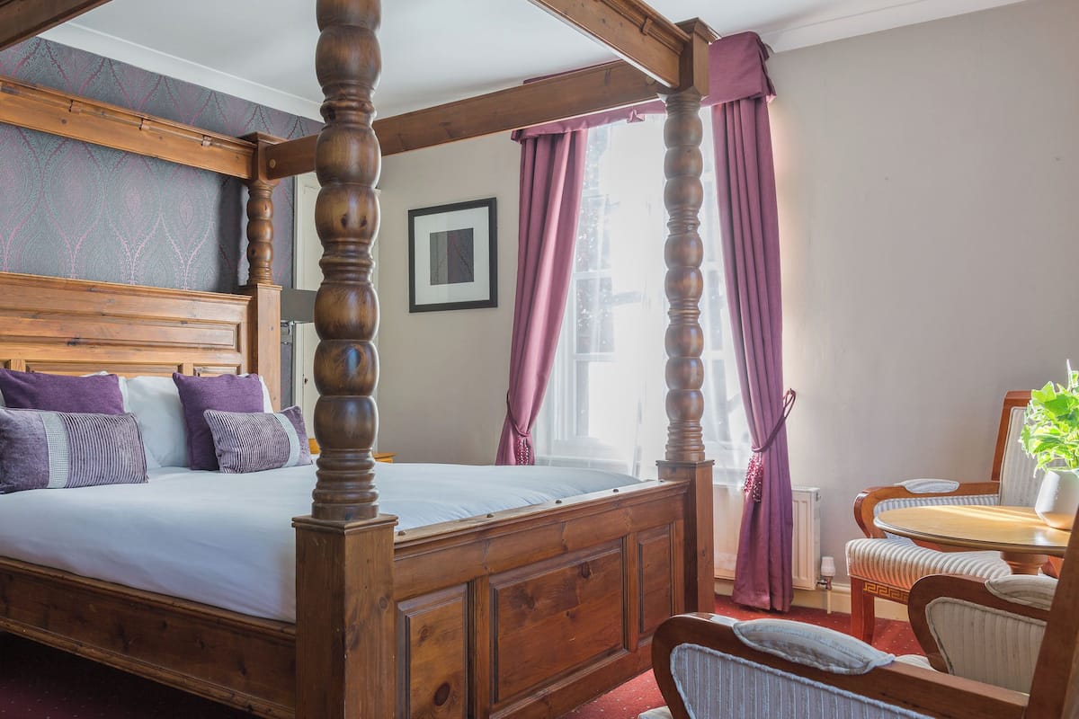 A four-poster bed with a wooden headboard, a bedside table, a chair, and a window with curtains.