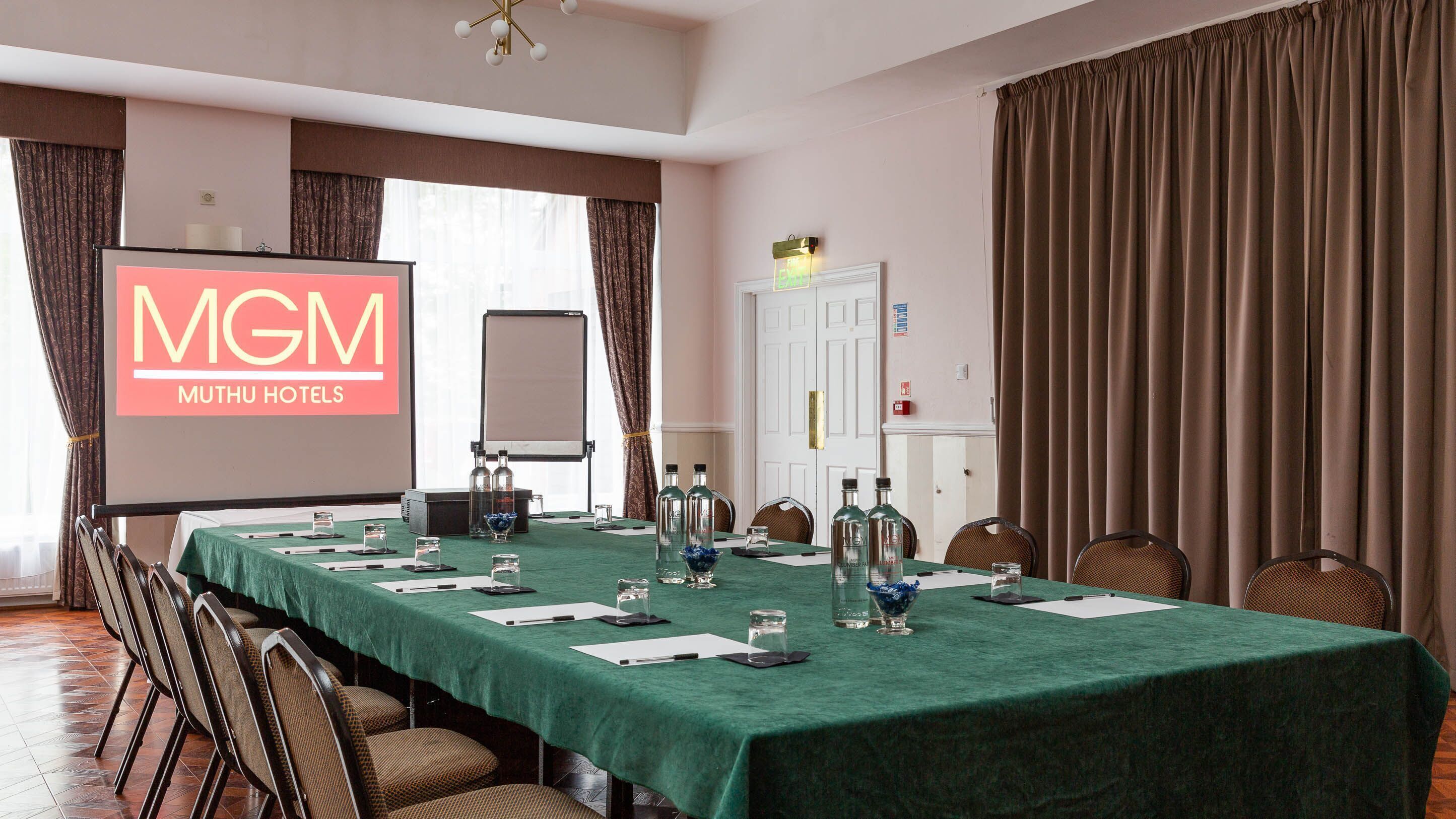Een congreszaal bij Muthu Hotels met een groene tafel, stoelen, een projectorscherm en een whiteboard.