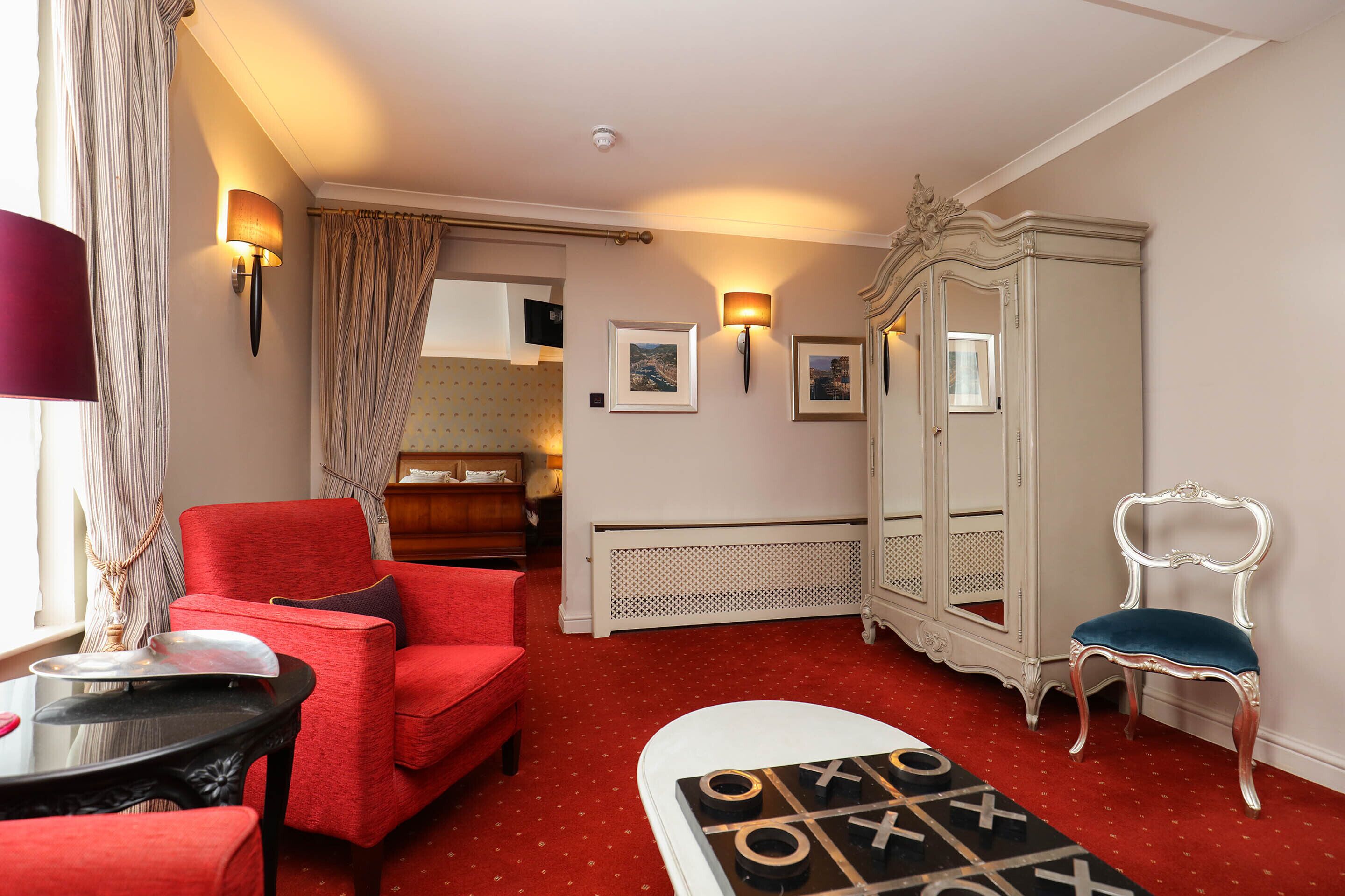 Ein Hotelzimmer mit rotem Teppich, einem roten Sessel, einem weißen Schrank, einem blauen Stuhl und einem Tisch für Tic-Tac-Toe.