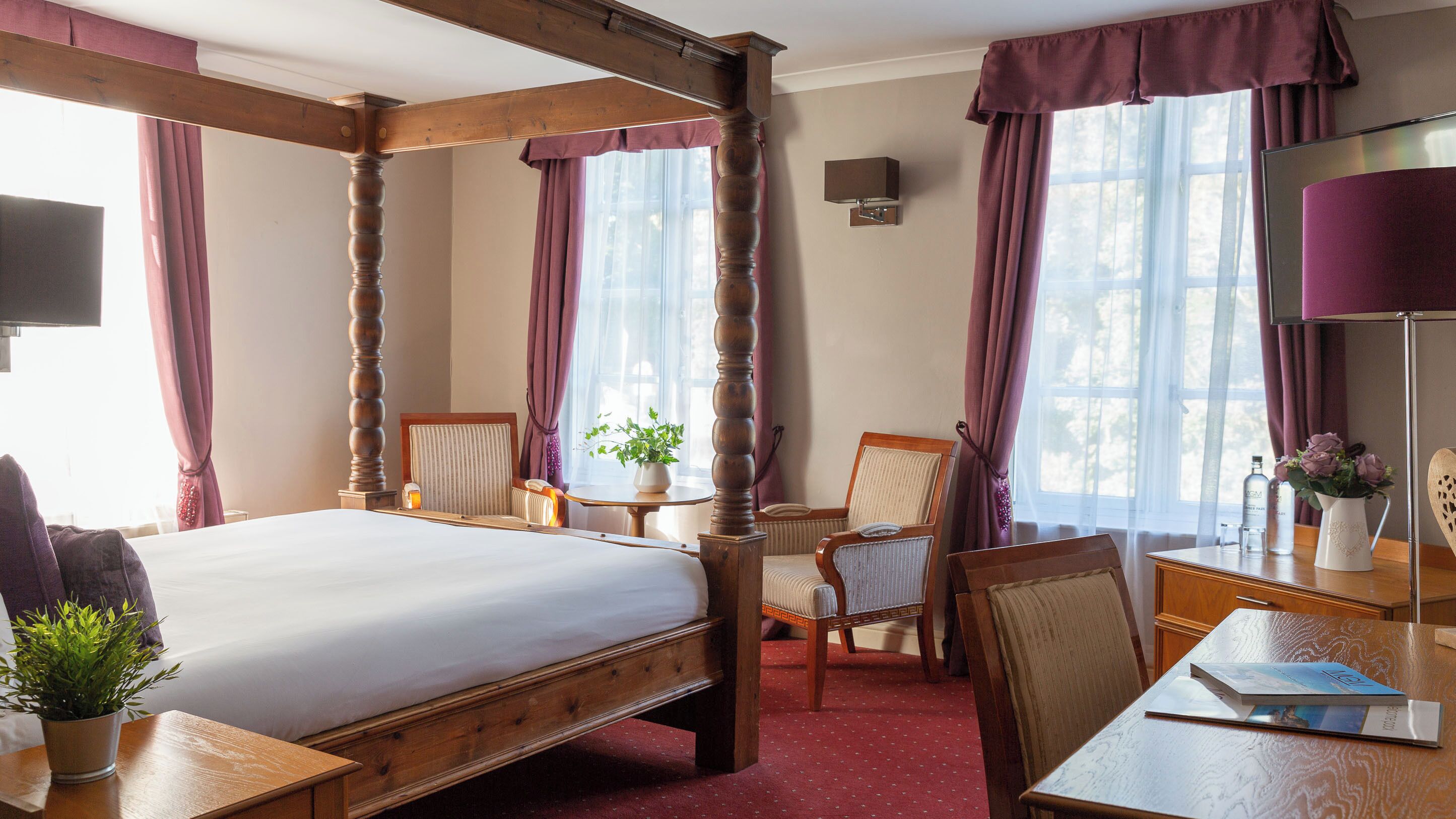 A four-poster bed with a wooden headboard, a bedside table, a chair, a desk, and a lamp.