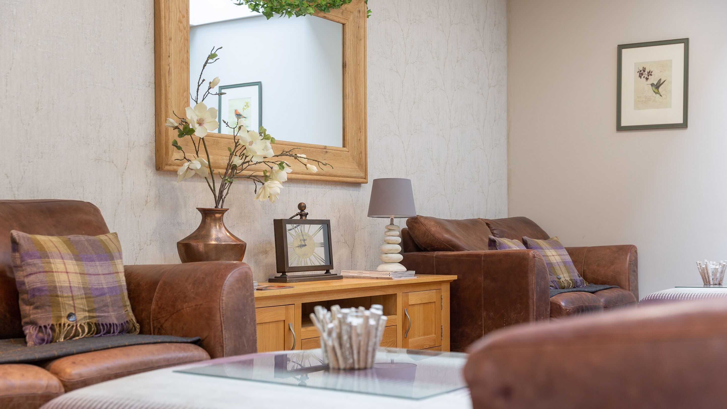 A living room with a brown leather sofa, a wooden side table with a lamp, a vase with flowers, and a framed picture on the wall.
