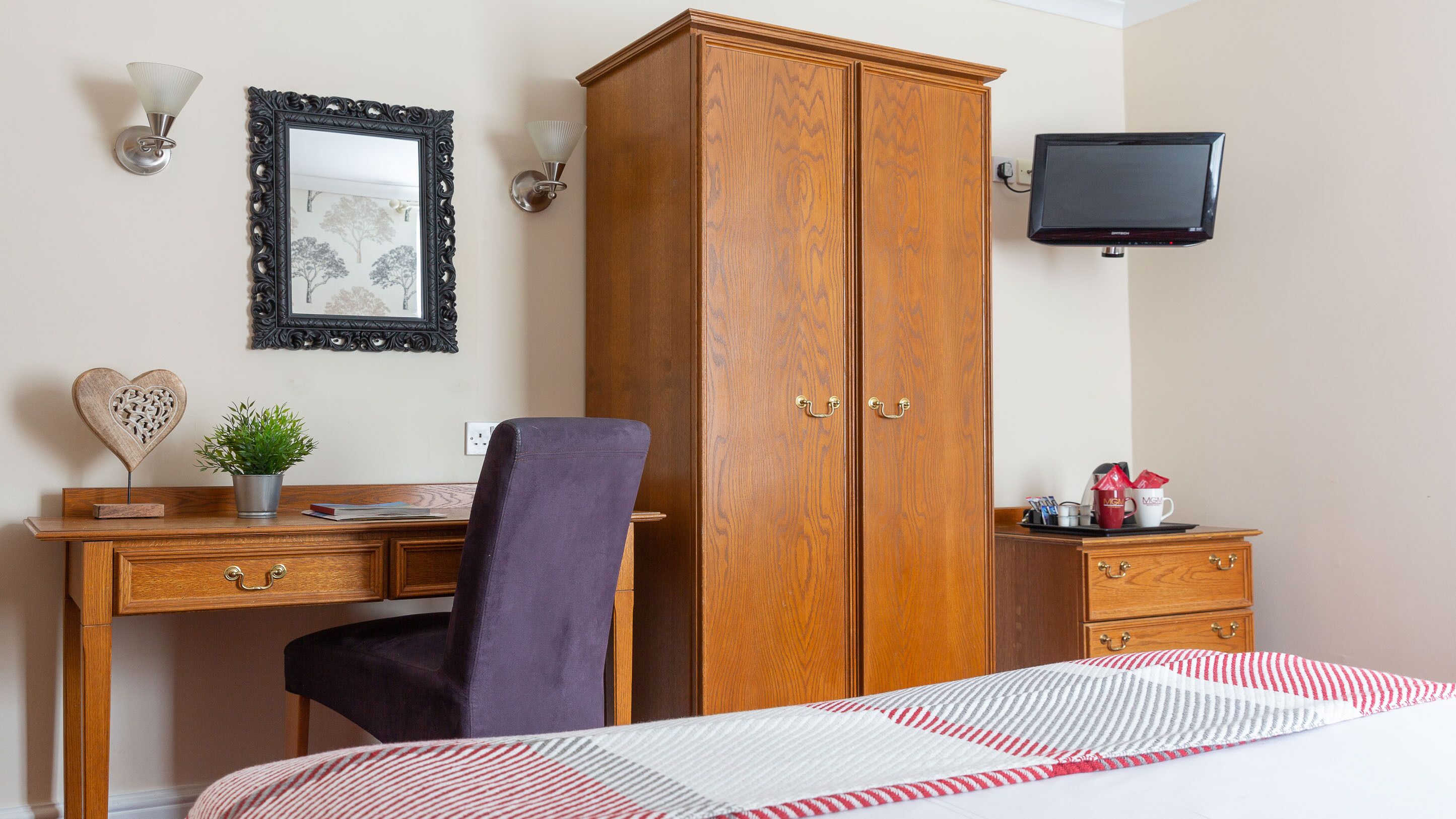 A hotel room with a wooden wardrobe, a desk with a chair, a bed with a striped bedspread, a television mounted on the wall, and a wall-mounted light fixture.