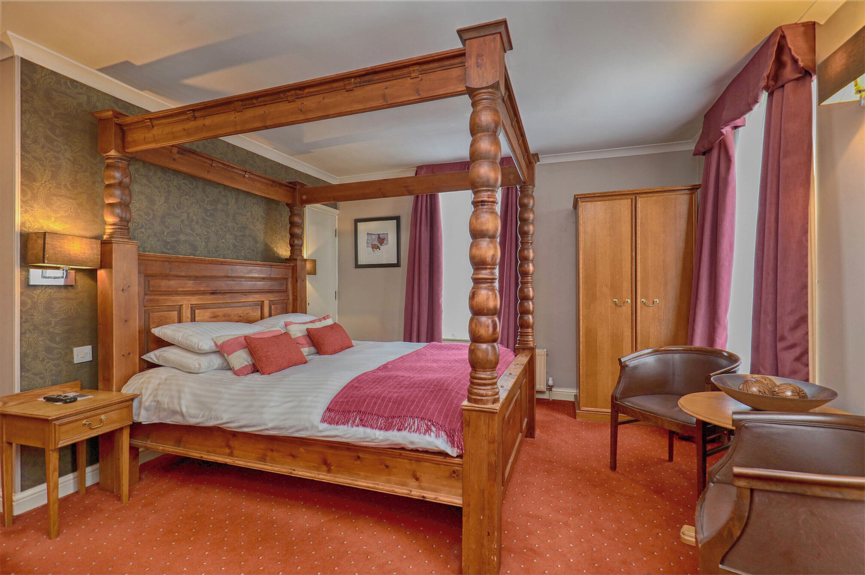 A four-poster bed with a wooden headboard, a bedside table, a chair, a small table with a bowl, and a wardrobe.