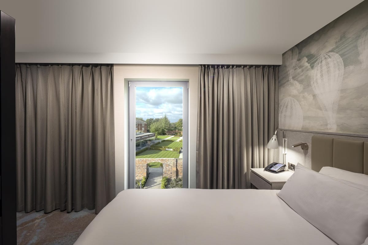 Une chambre d’hôtel avec un lit, une table de chevet avec une lampe et une vue sur un jardin, visible par une porte.