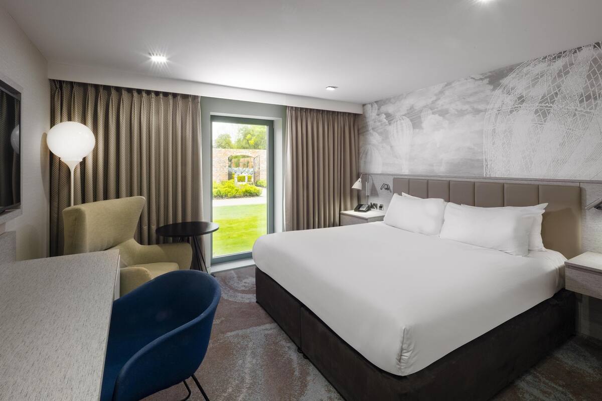 Une chambre d’hôtel avec un grand lit, un bureau, une chaise, une petite table et une vue sur un jardin, visible par la porte.