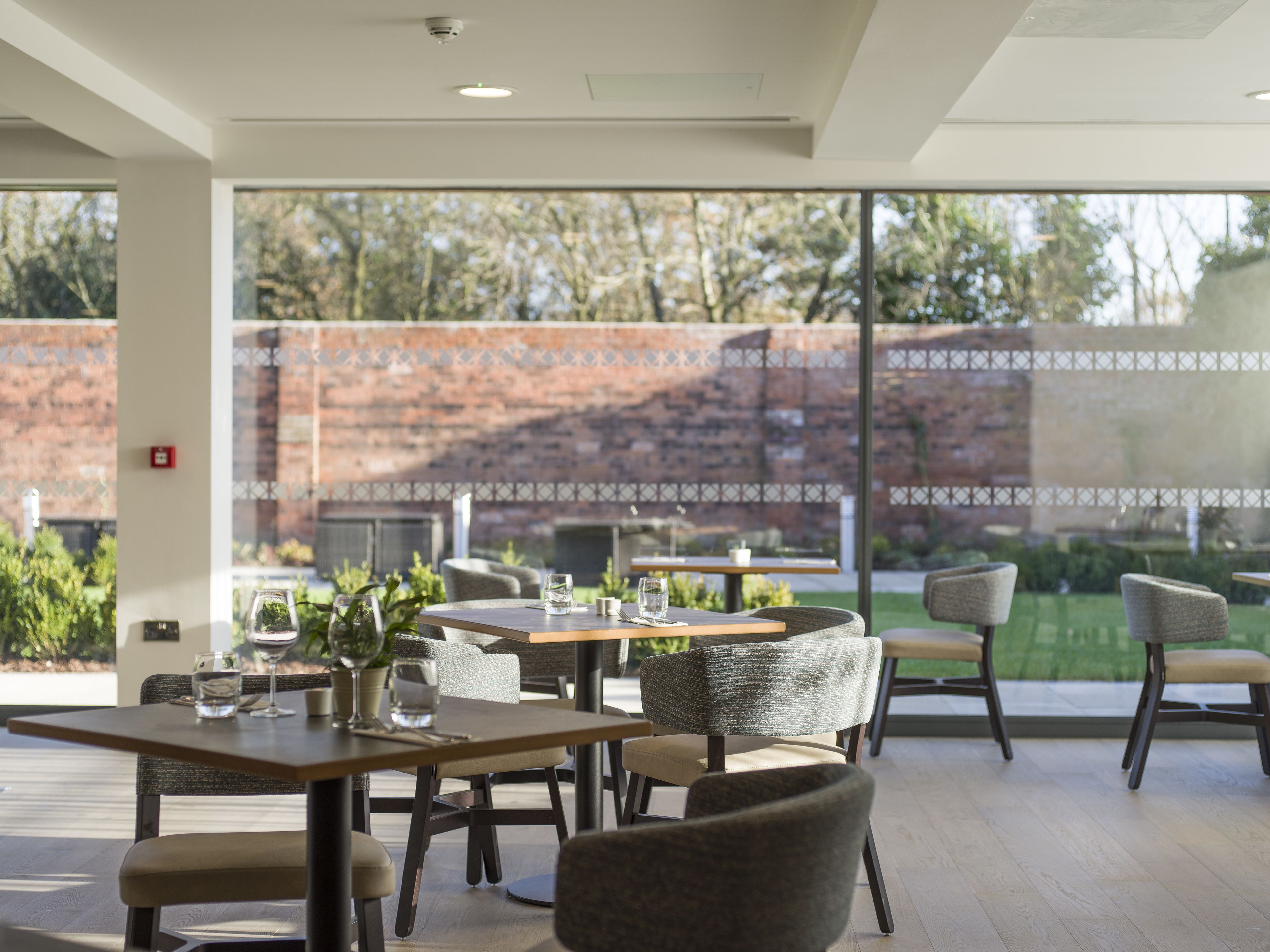 A modern outdoor dining area with tables and chairs, large windows offering a view of a brick wall and greenery.