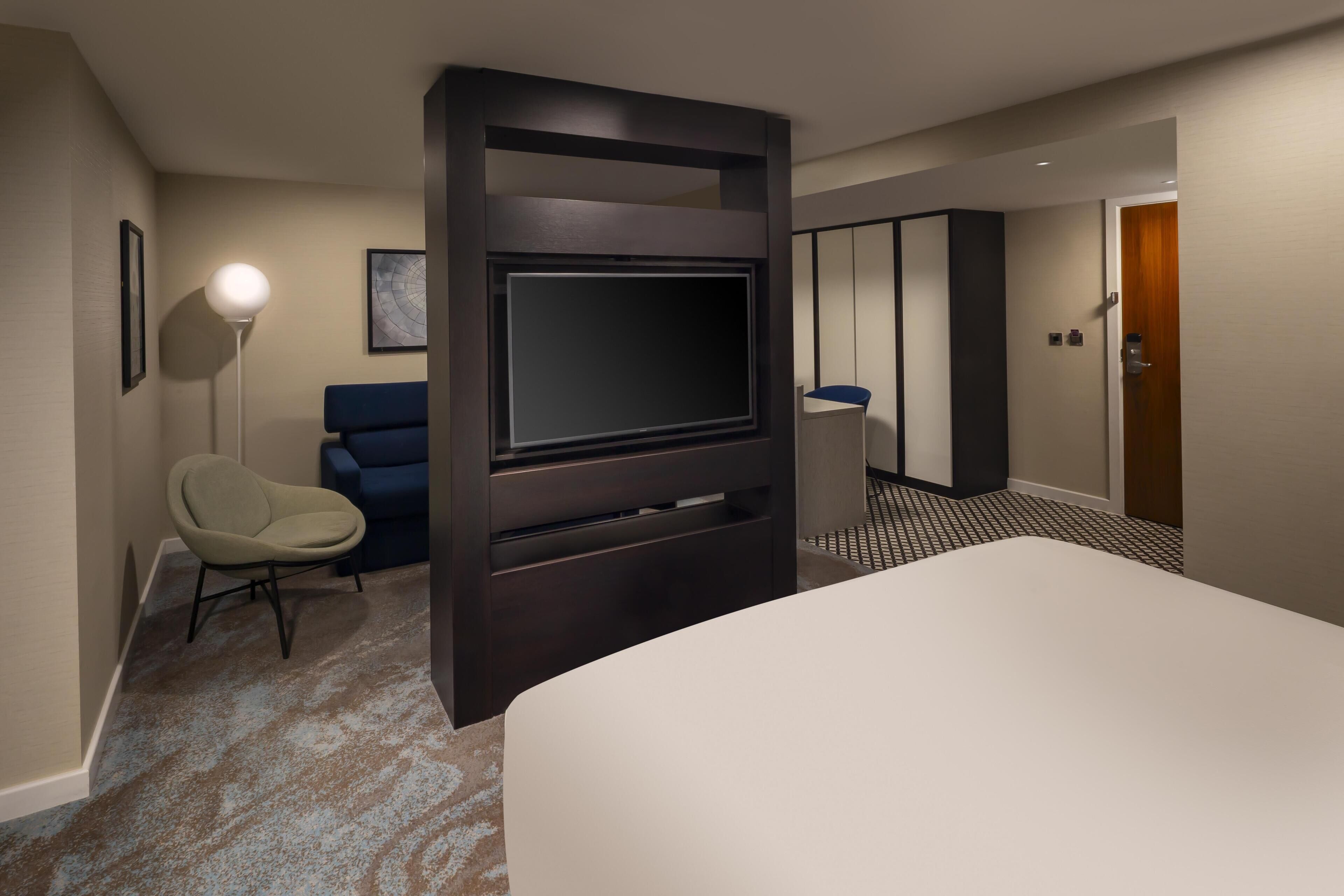 A hotel room with a large bed, a TV mounted on a dark wall, a desk with a chair, and a lamp.