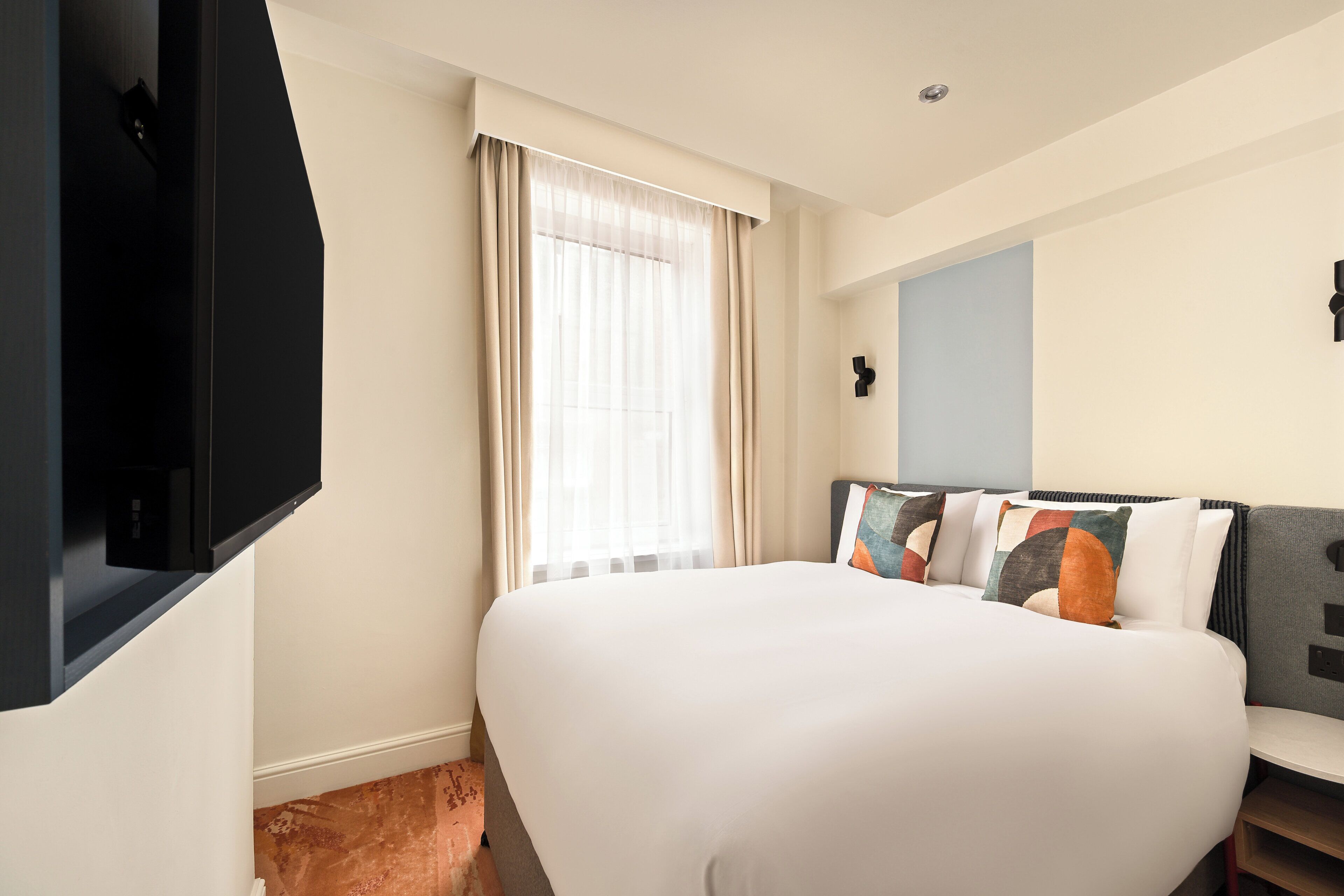 Une chambre d’hôtel comprenant un lit, une télévision, une fenêtre avec des rideaux et un luminaire fixé au mur.