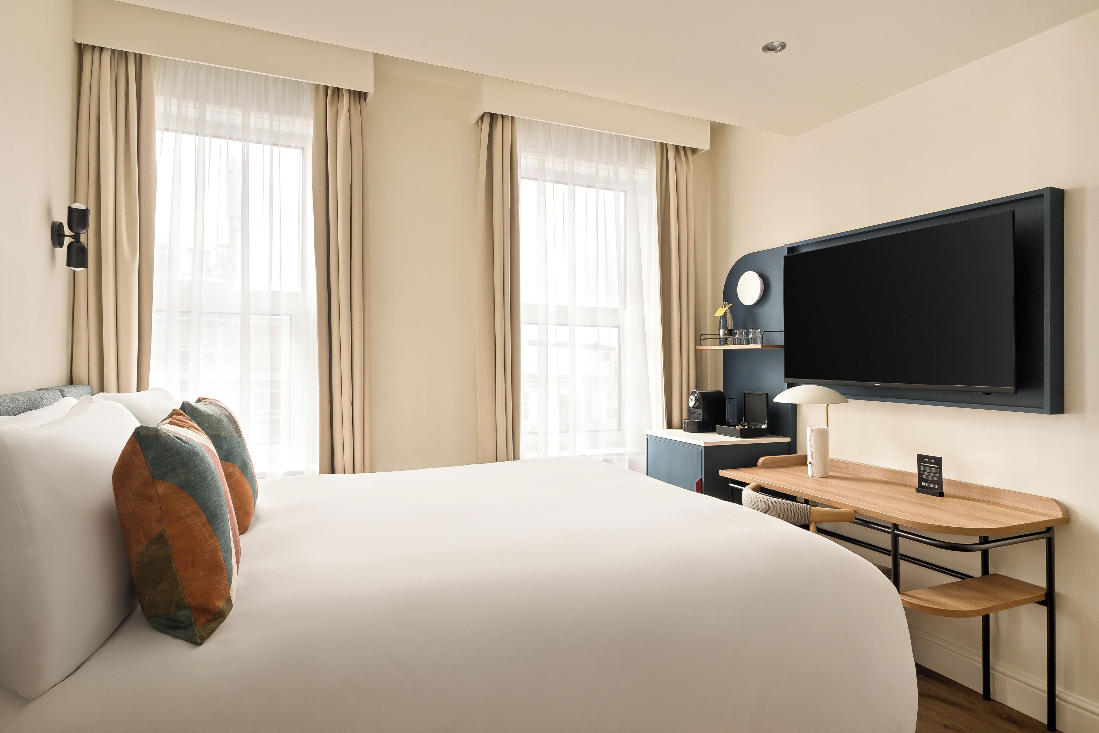 A hotel room with a large bed, a desk with a lamp, a flat-screen TV, and a window with curtains.