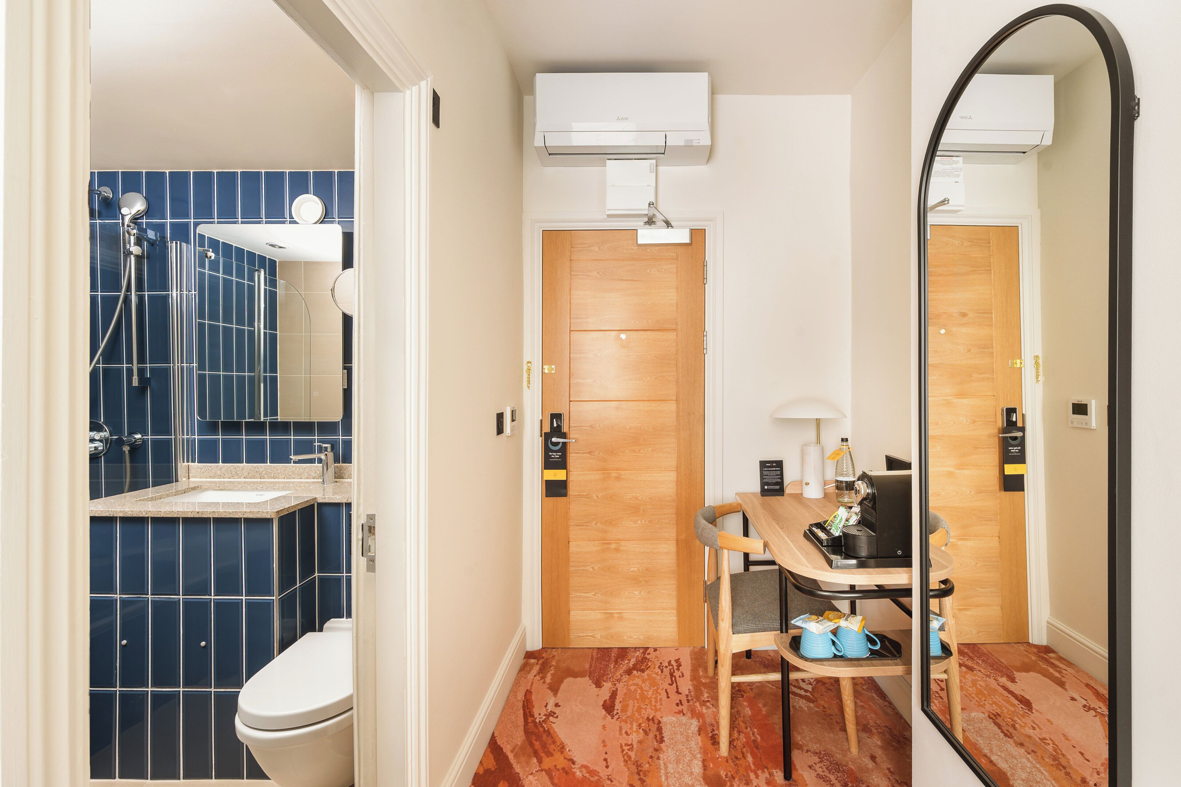 Un baño con ducha de azulejos azules, inodoro blanco, puerta de madera y un espejo que refleja la habitación.