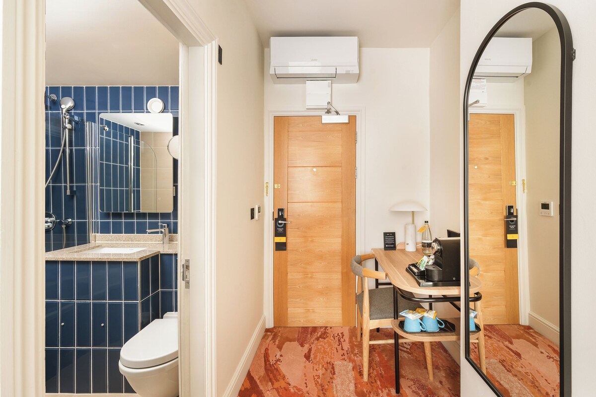 A bathroom with a blue-tiled shower, a white toilet, a wooden door, and a mirror reflecting the room.
