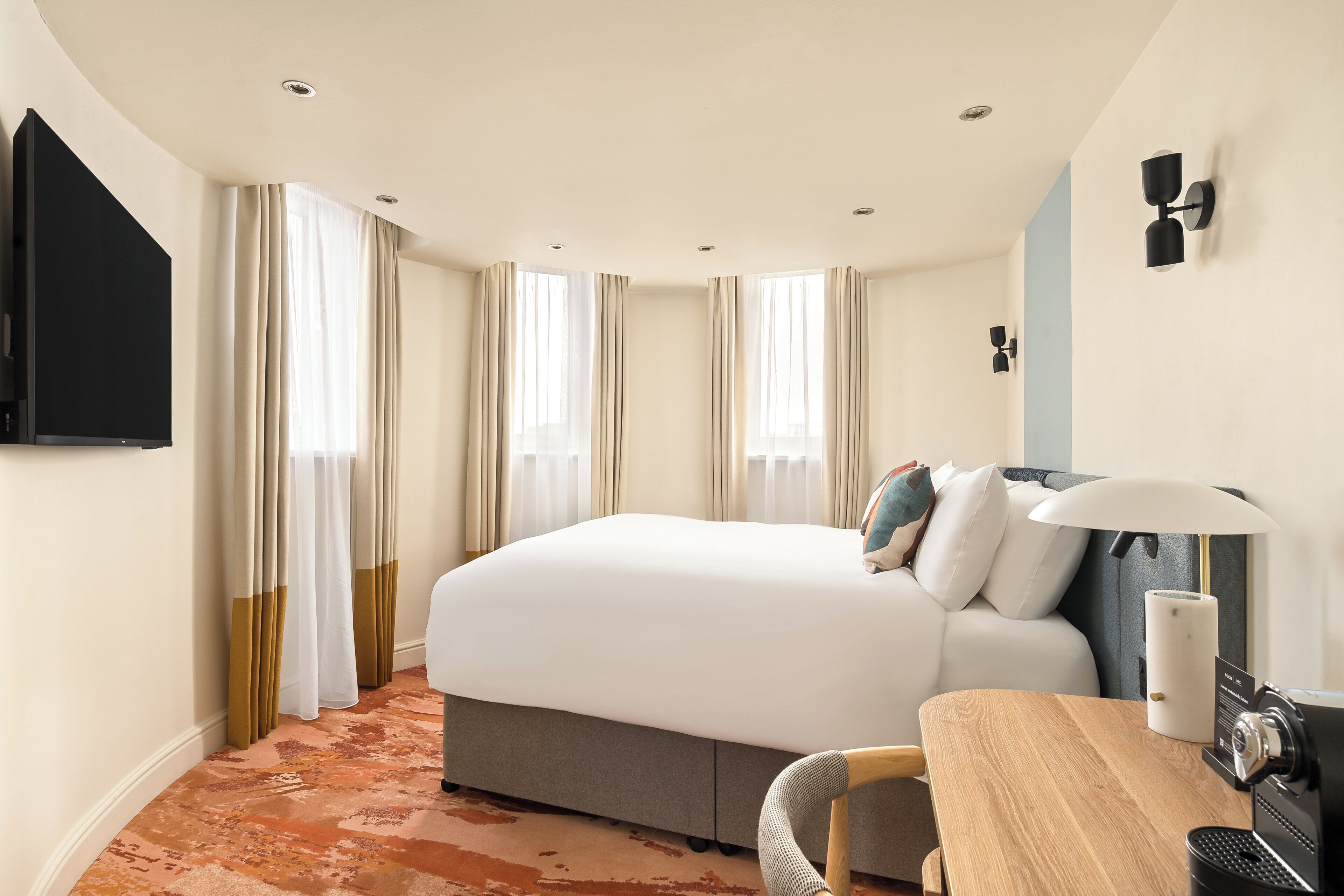 Una habitación de hotel con una cama, un escritorio con lámpara, un televisor y una ventana con cortinas.
