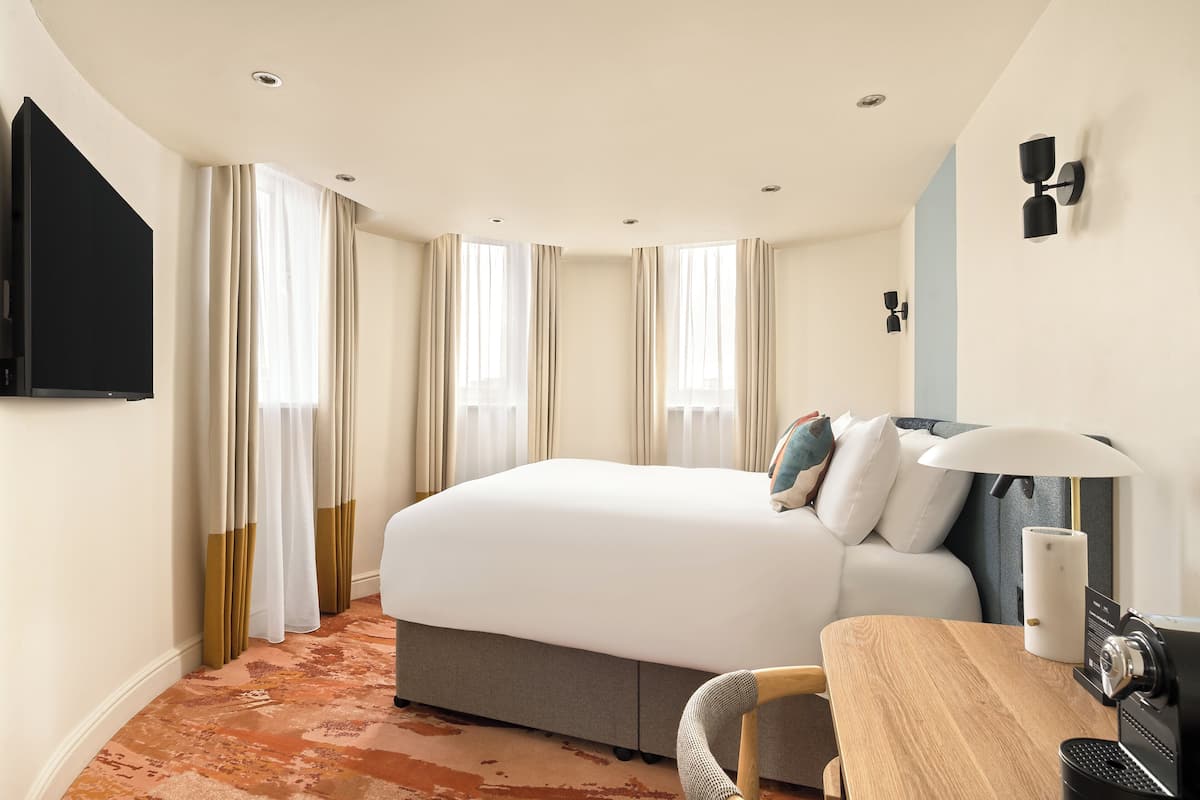 A hotel room with a bed, a desk with a lamp, a television, and a window with curtains.