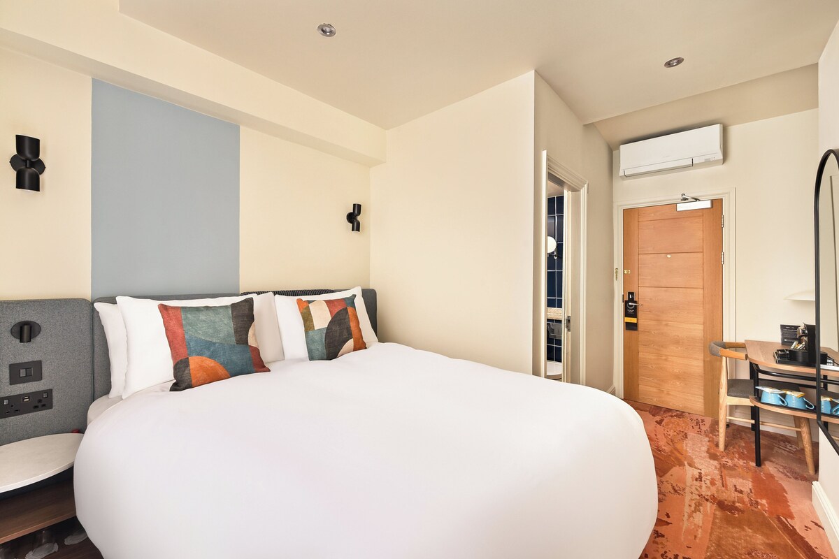 A hotel room with a large bed, a desk with a chair, a wardrobe, and an air conditioning unit.