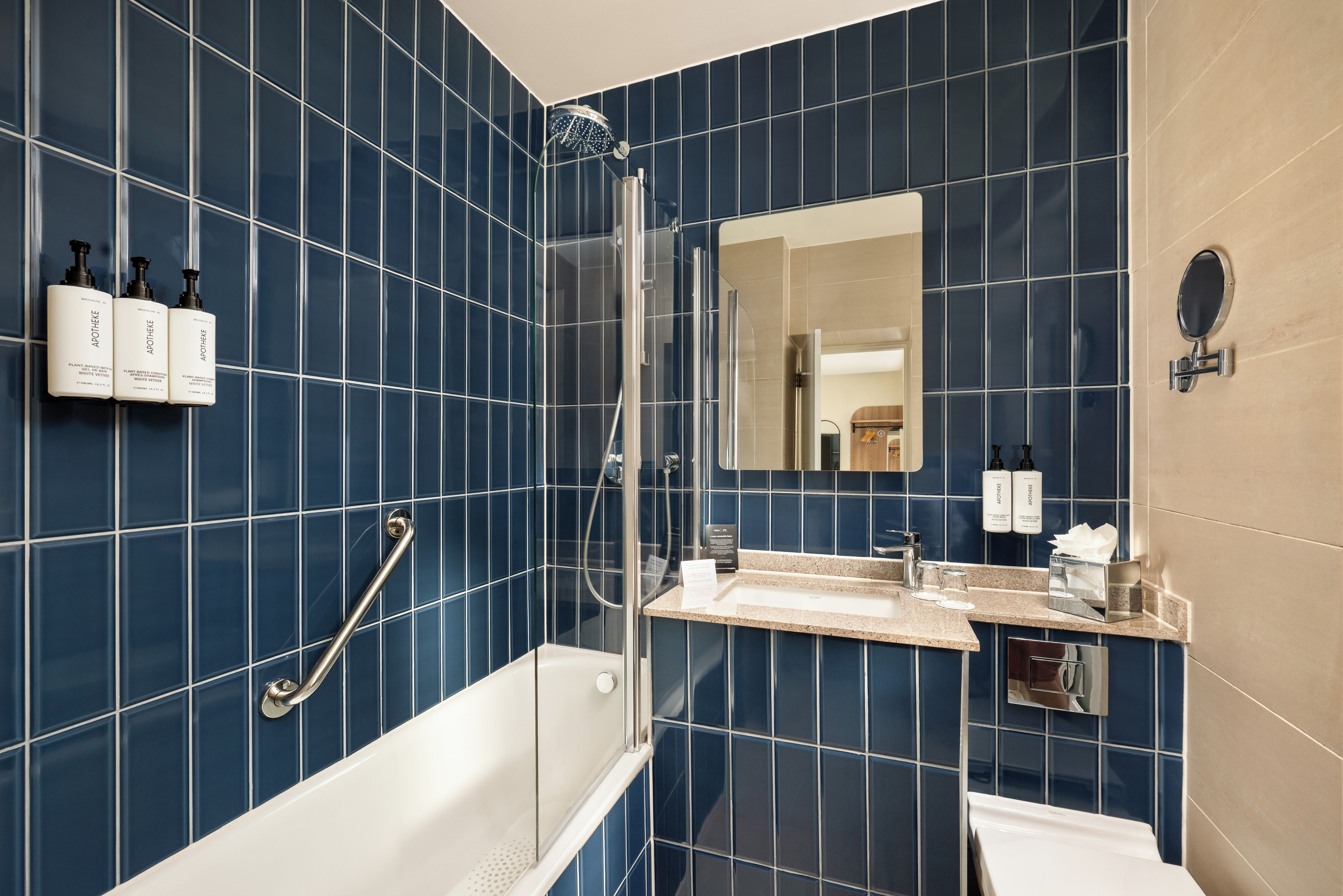A modern bathroom with blue tiles, a glass-enclosed shower, a white bathtub, a vanity with a mirror, and toiletries on the wall.