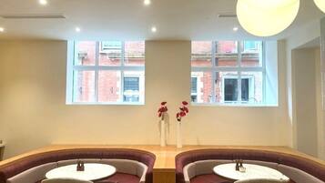 A seating area with cushioned benches and round tables, large windows with a view of buildings, and a central vase with red flowers.