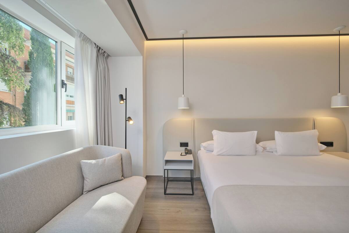 A modern hotel room with a large bed, a sofa, a bedside table with a lamp, and a view of the outside.