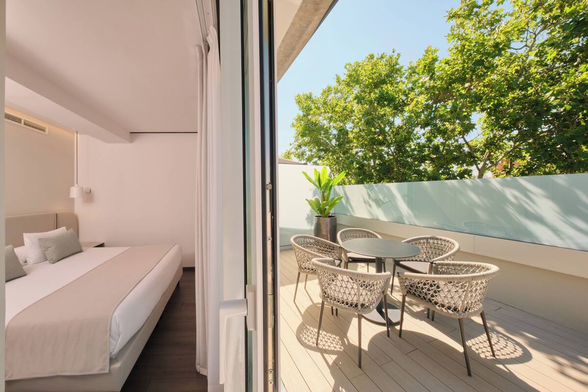 A modern bedroom with a bed, bedside table, and a view of a balcony with wicker chairs and a potted plant.