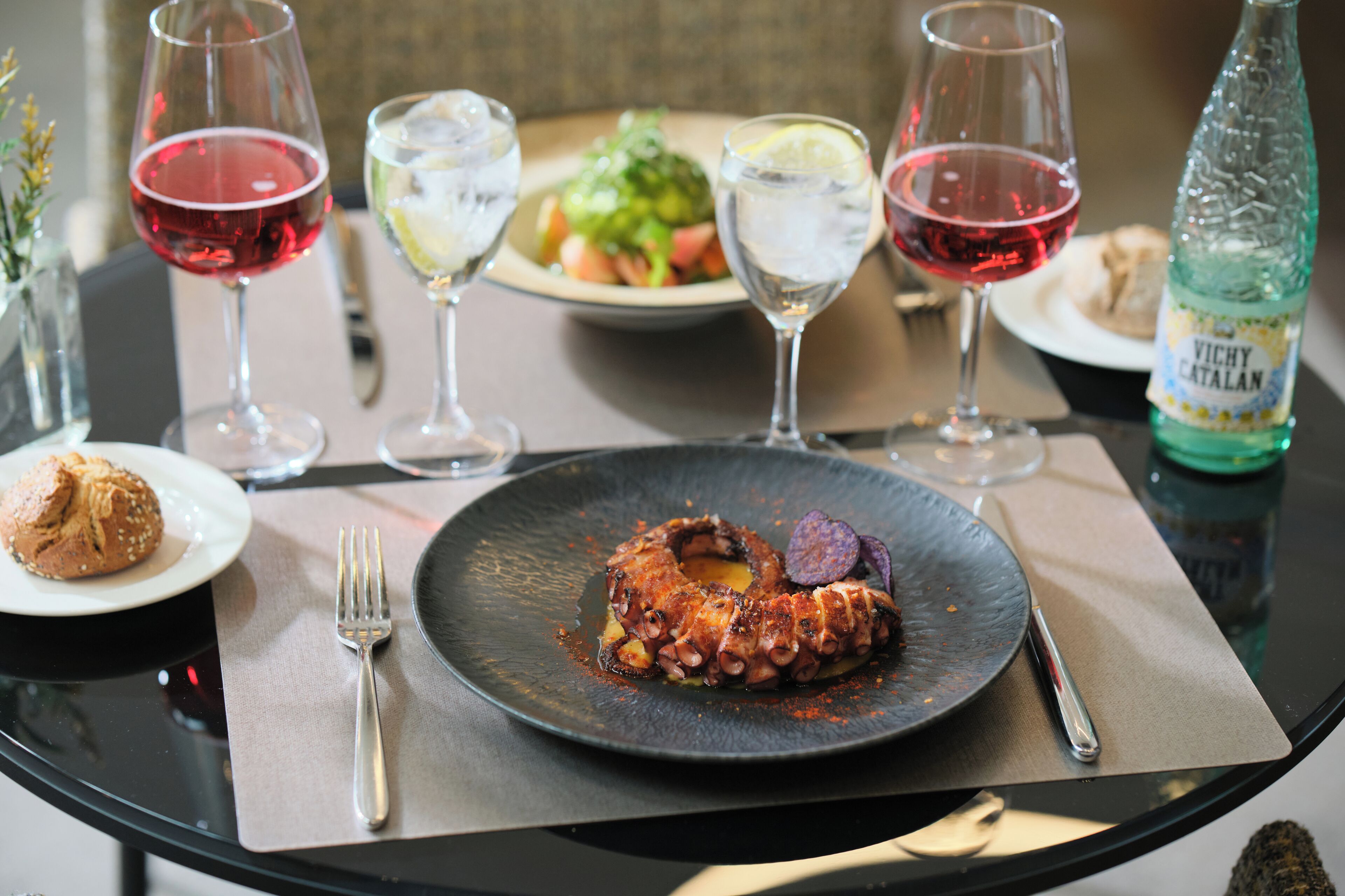 A dining table with a plate of octopus, wine glasses, a bottle of Vichy Catalan water, and a muffin.