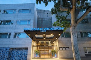 The exterior of the 1881 Madrid Ventas Hotel with a modern facade, large glass entrance, and a tree in the foreground.