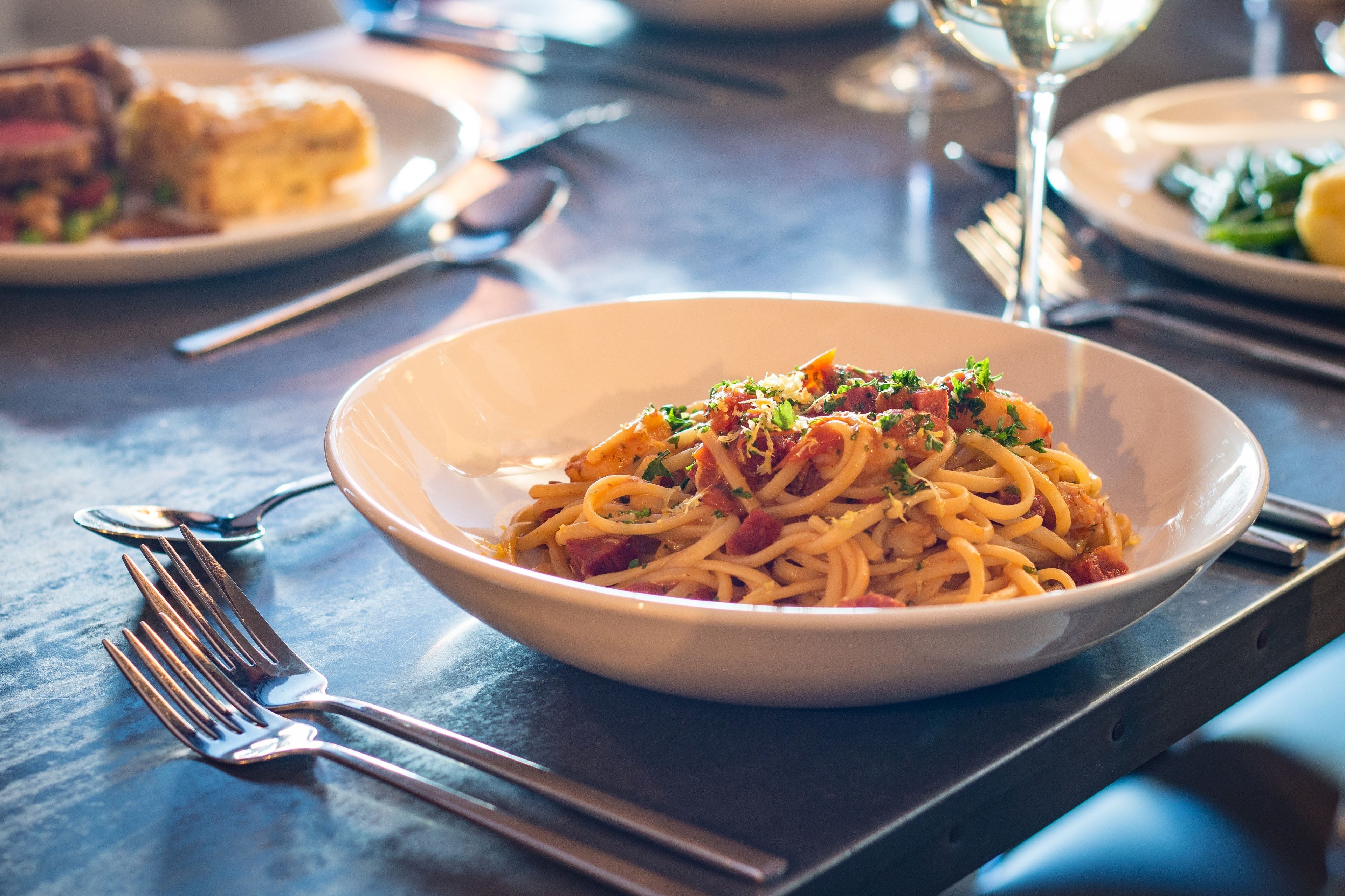 A dining table with a bowl of pasta, cutlery, and a glass of wine.