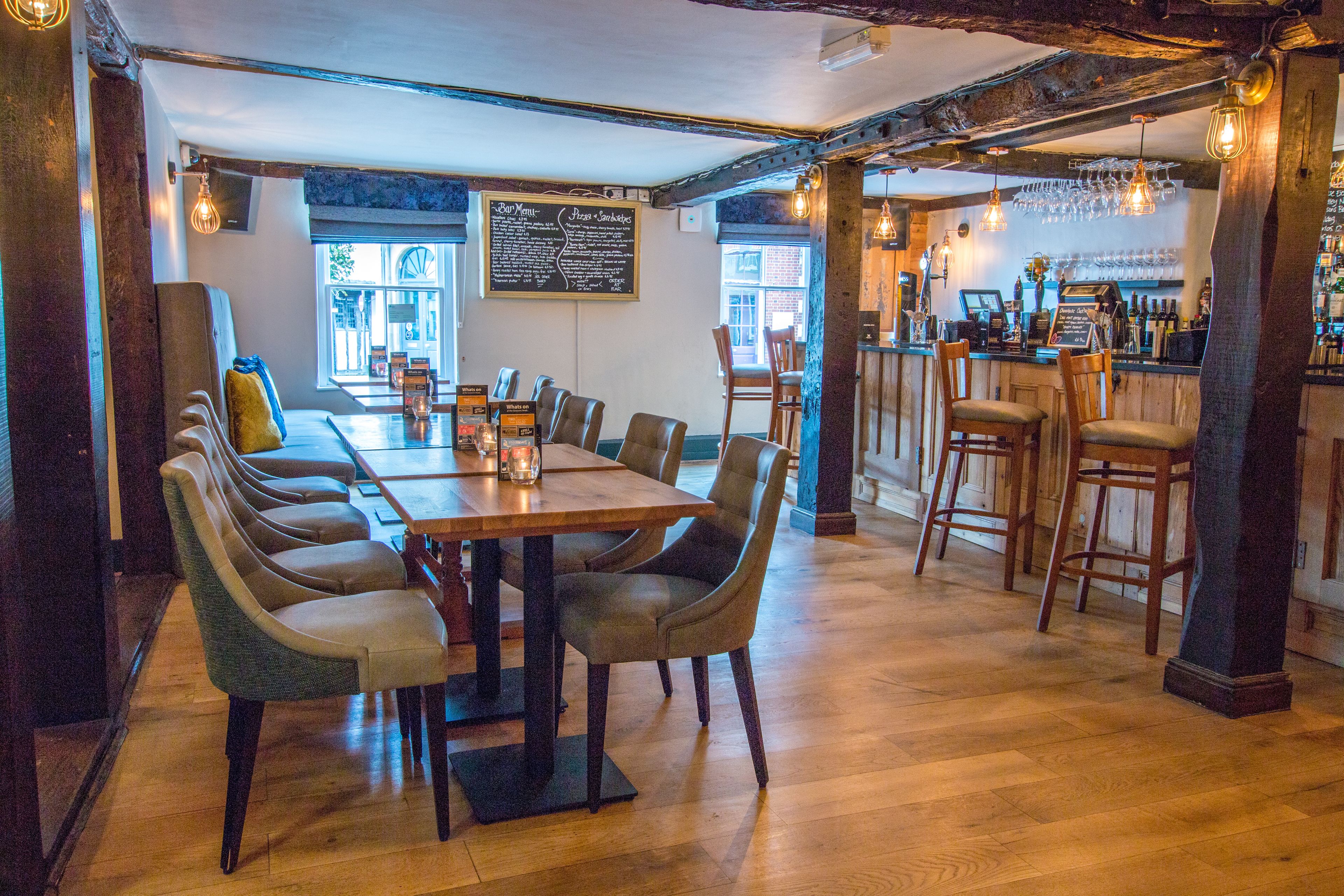 Un restaurante con una larga mesa de madera y varias sillas, una zona de bar con taburetes y un menú en una pizarra en la pared.