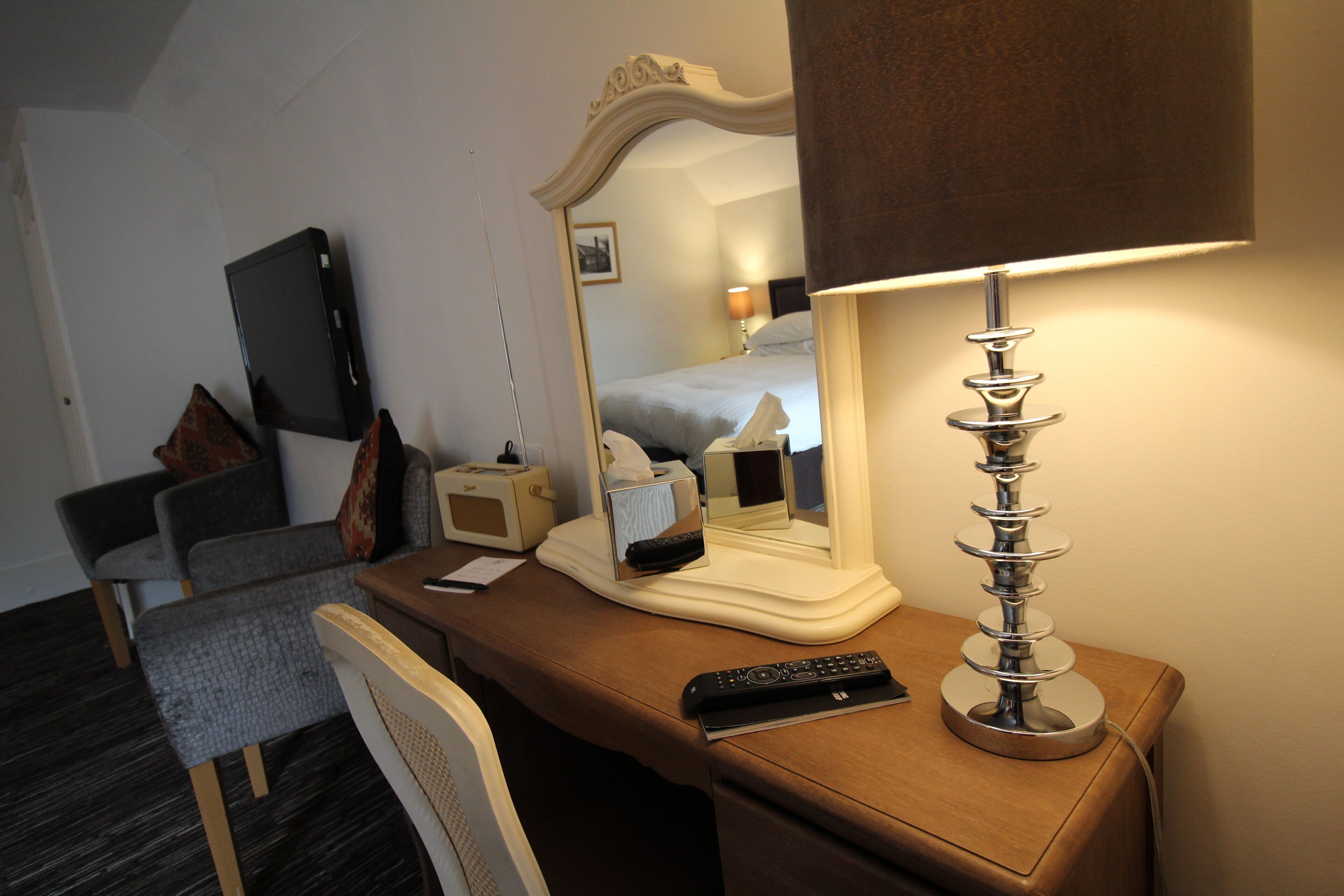 A hotel room with a desk, a chair, a bed, a mirror, a lamp, and a television.