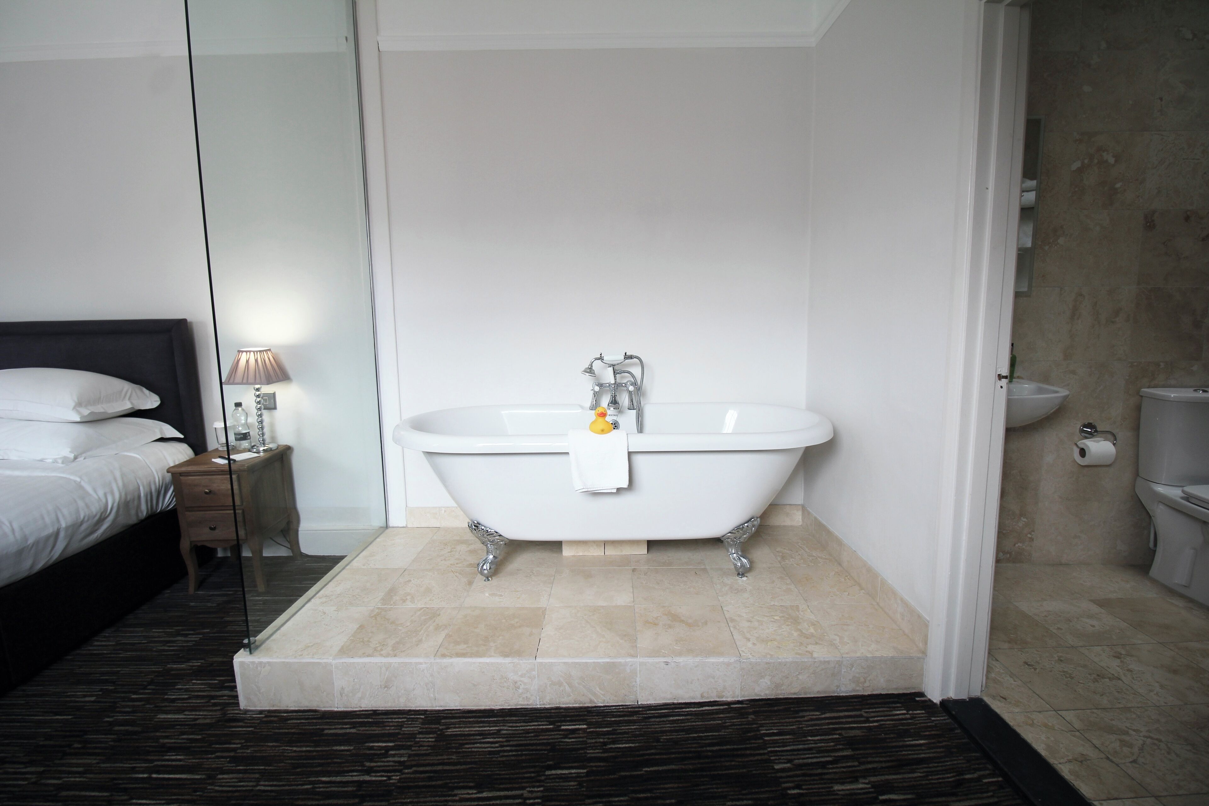 A modern bathroom with a freestanding bathtub, a bedside table with a lamp, and a toilet.