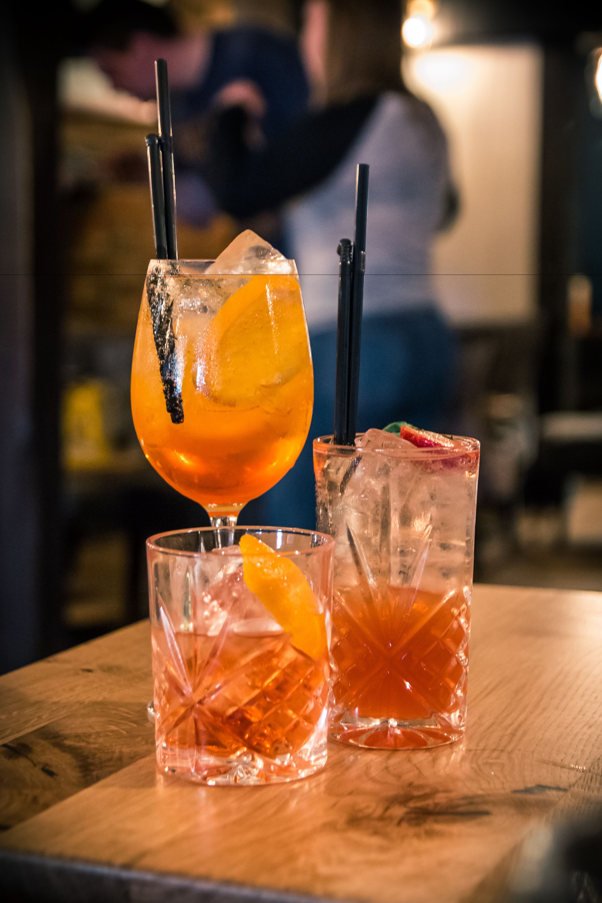 Três copos com bebidas alaranjadas, gelo e guarnições sobre uma mesa de madeira.