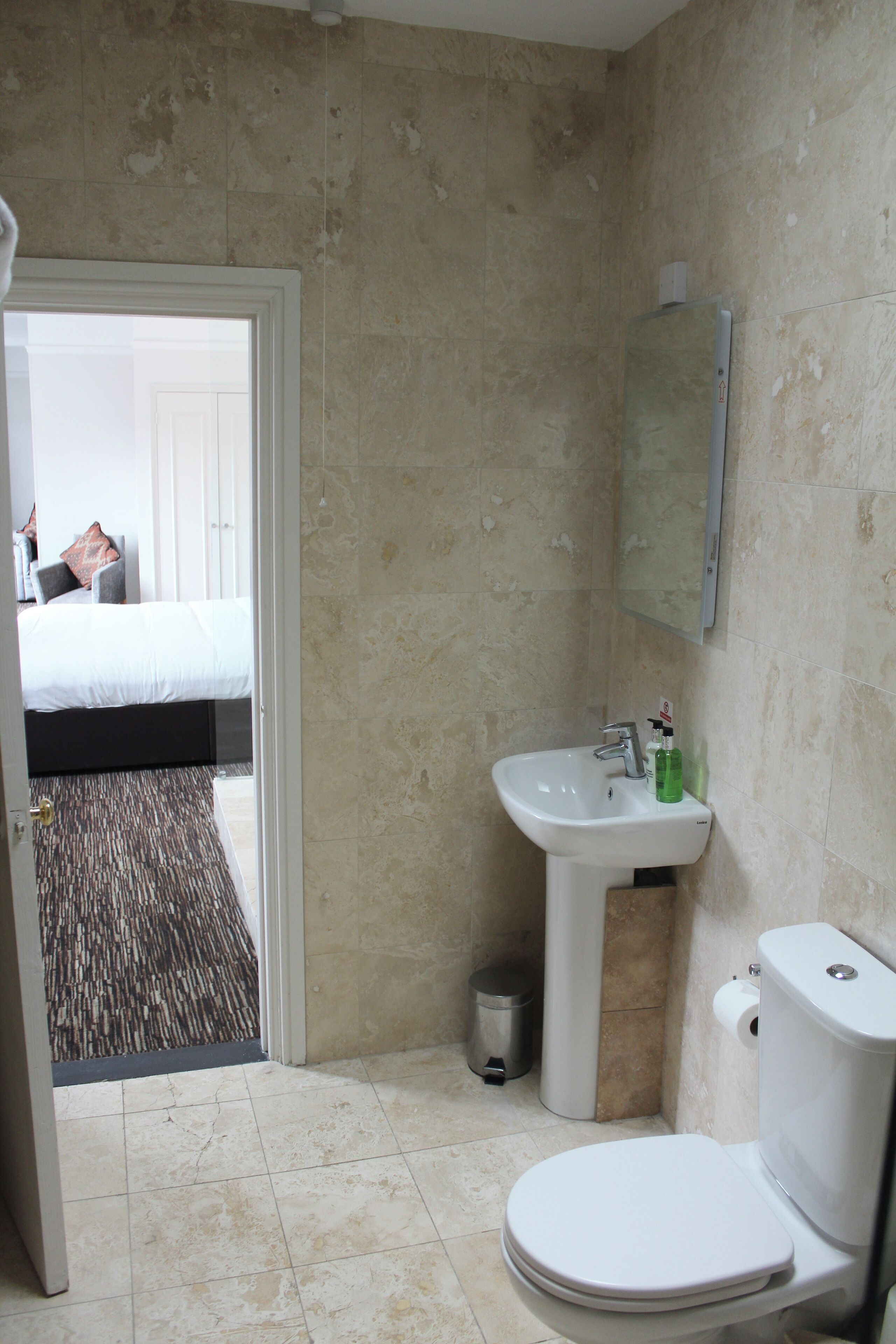 A bathroom with a toilet, sink, and mirror. There is a door leading to a bedroom with a bed and a view of the outside.