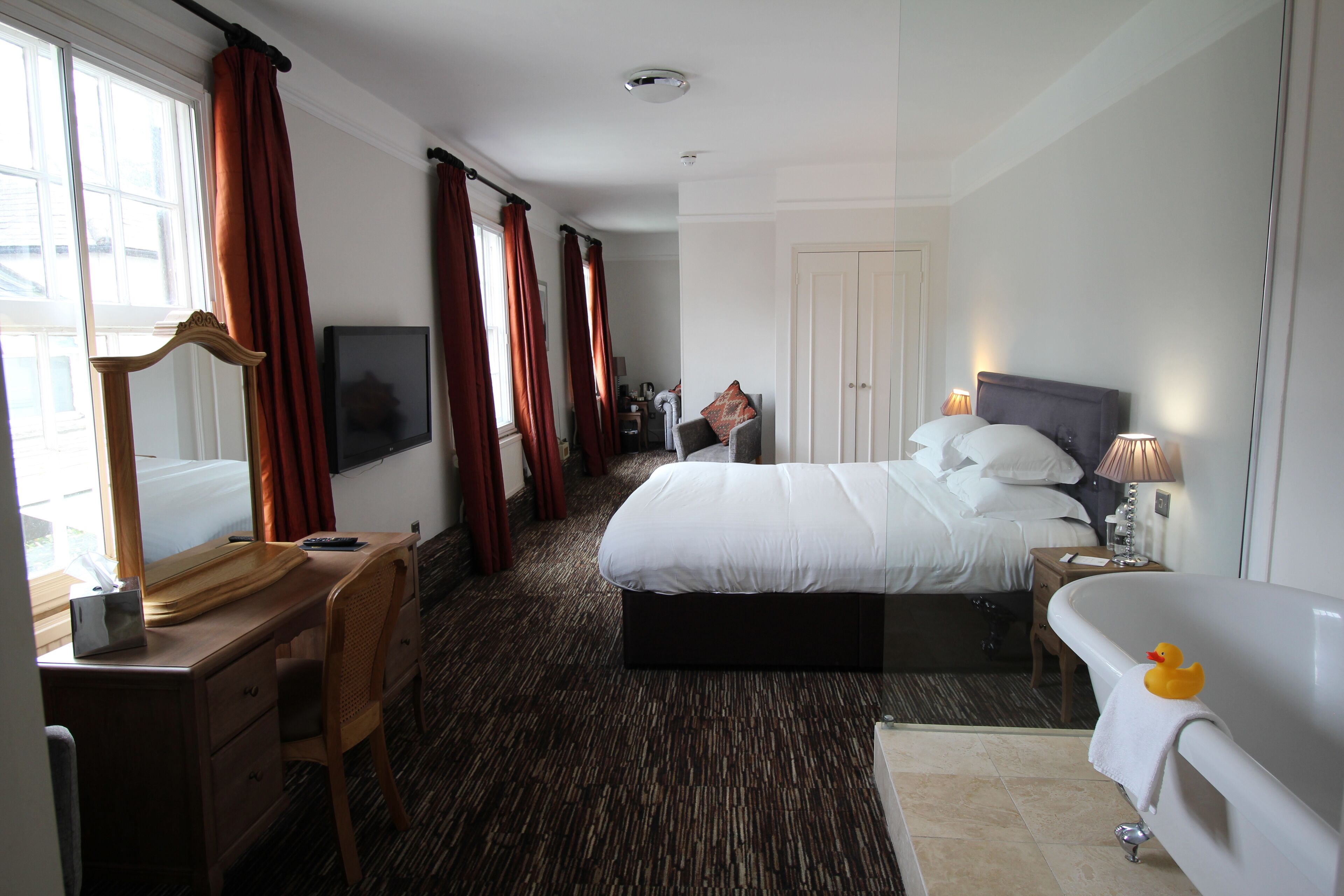 A hotel room with a bed, a desk with a chair, a desk lamp, a bedside table with a lamp, a bathtub with a rubber duck, and a large mirror.