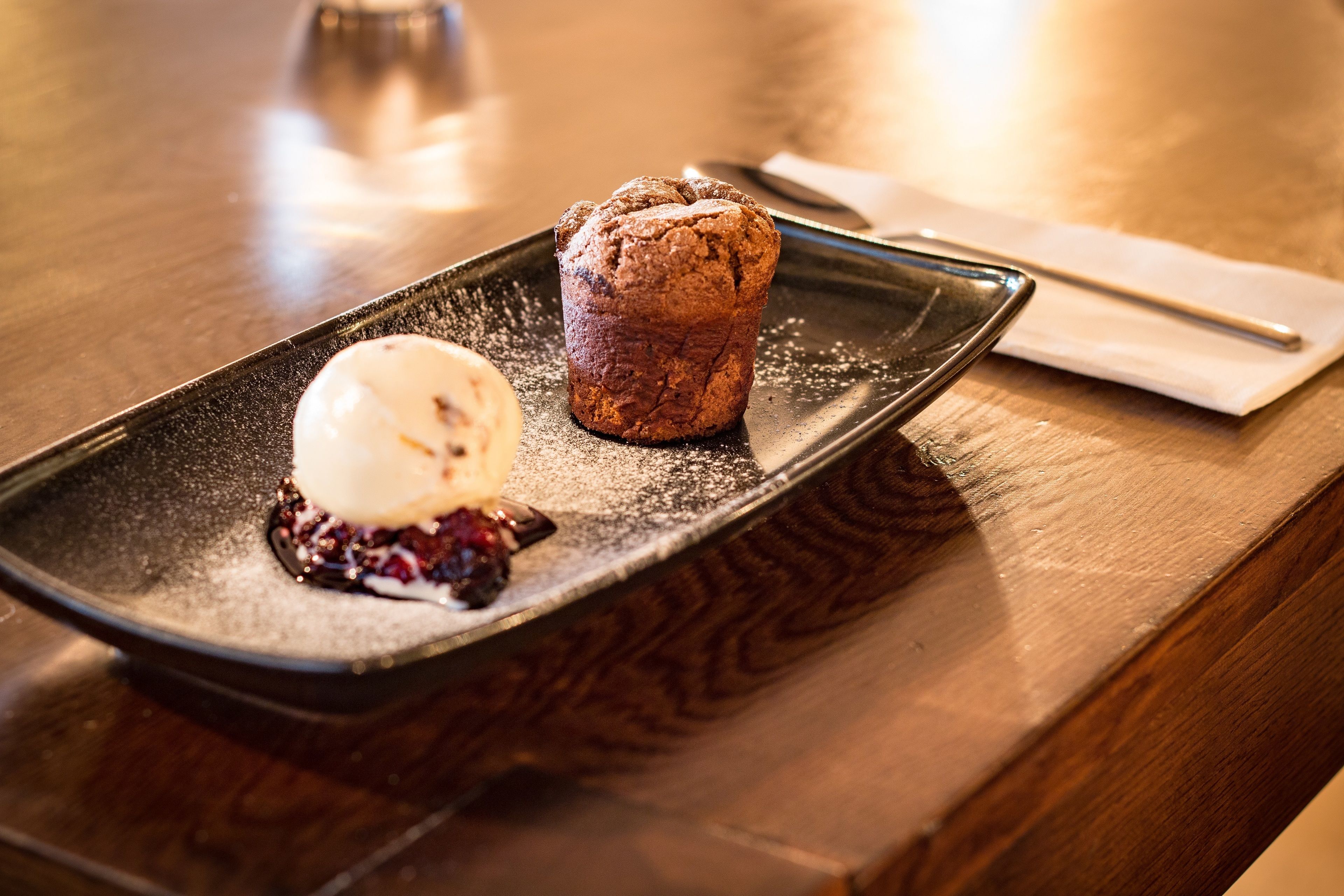 Un postre sobre un plato negro con un muffin y una bola de helado.