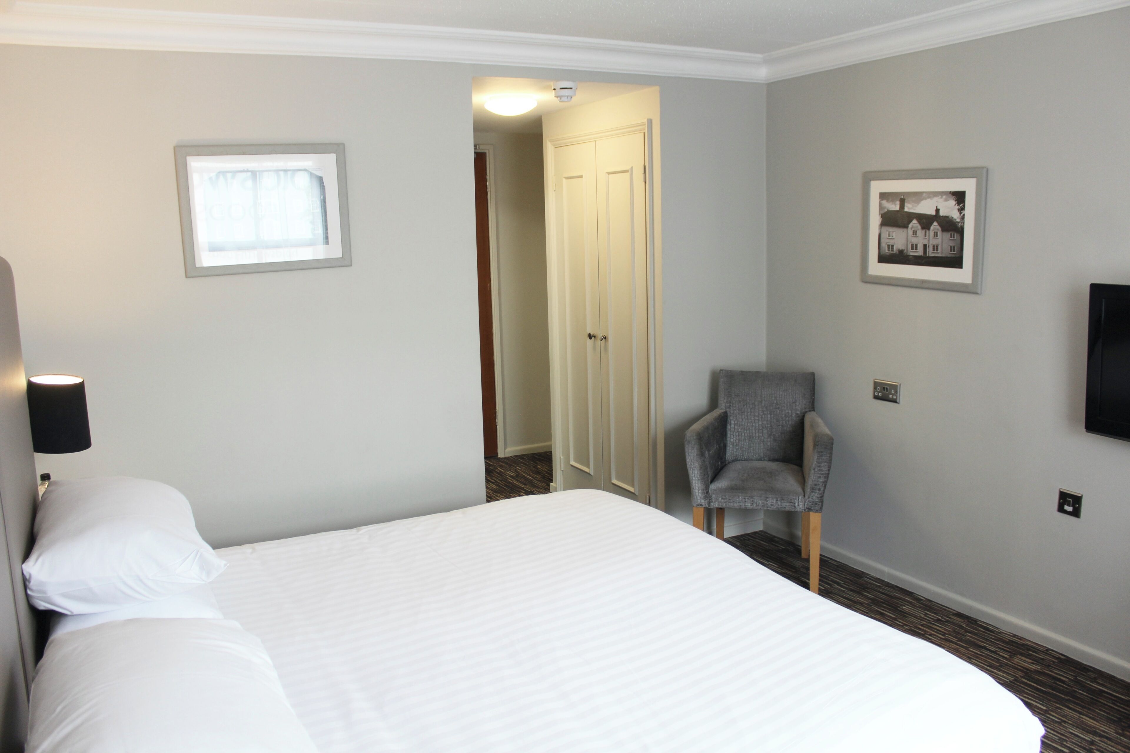 Een hotelkamer met een bed, een stoel, een ingelijst beeld en een deur naar een andere kamer.