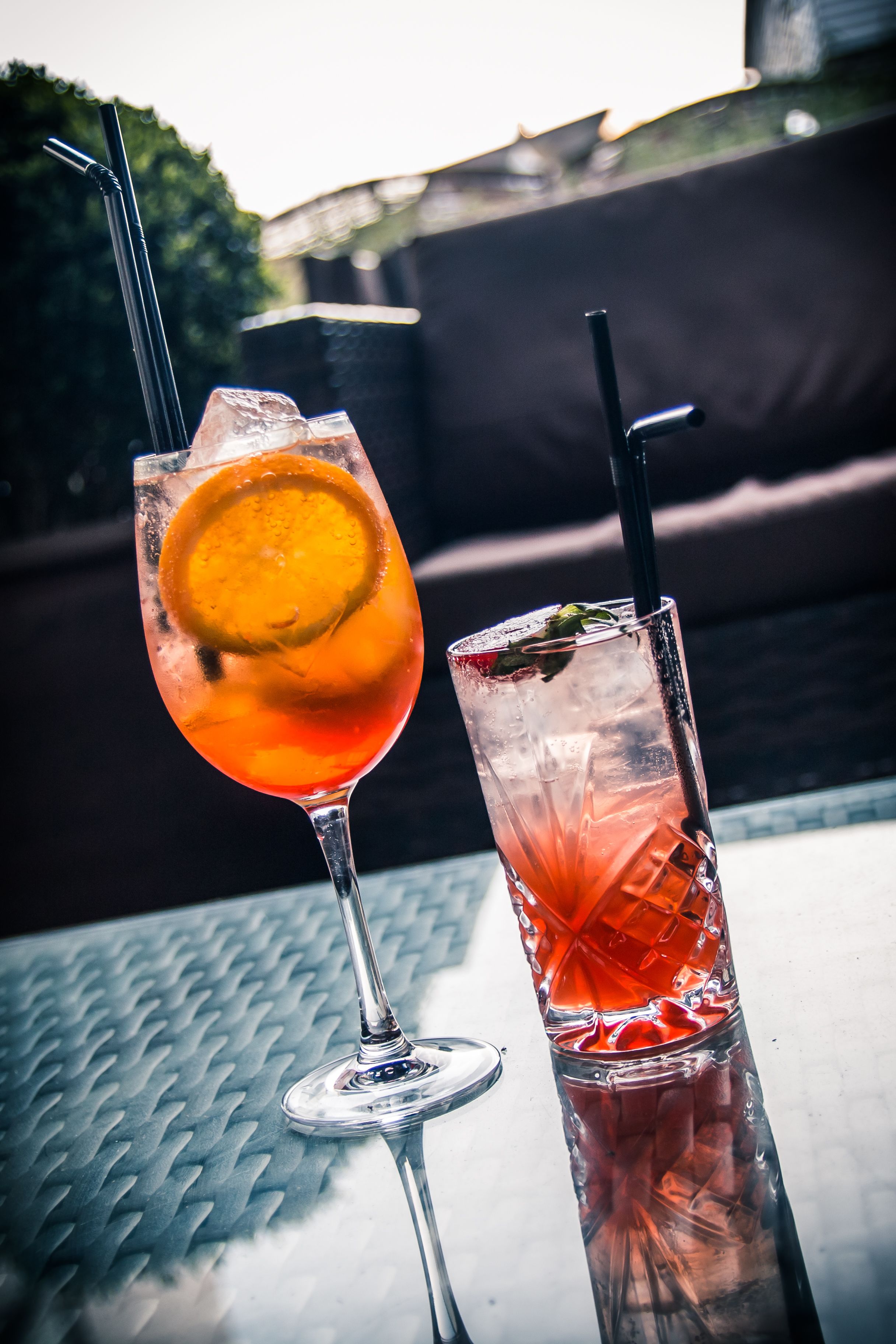 Two drinks on a table with a metal pattern, one with a lemon slice and the other with a mint garnish.