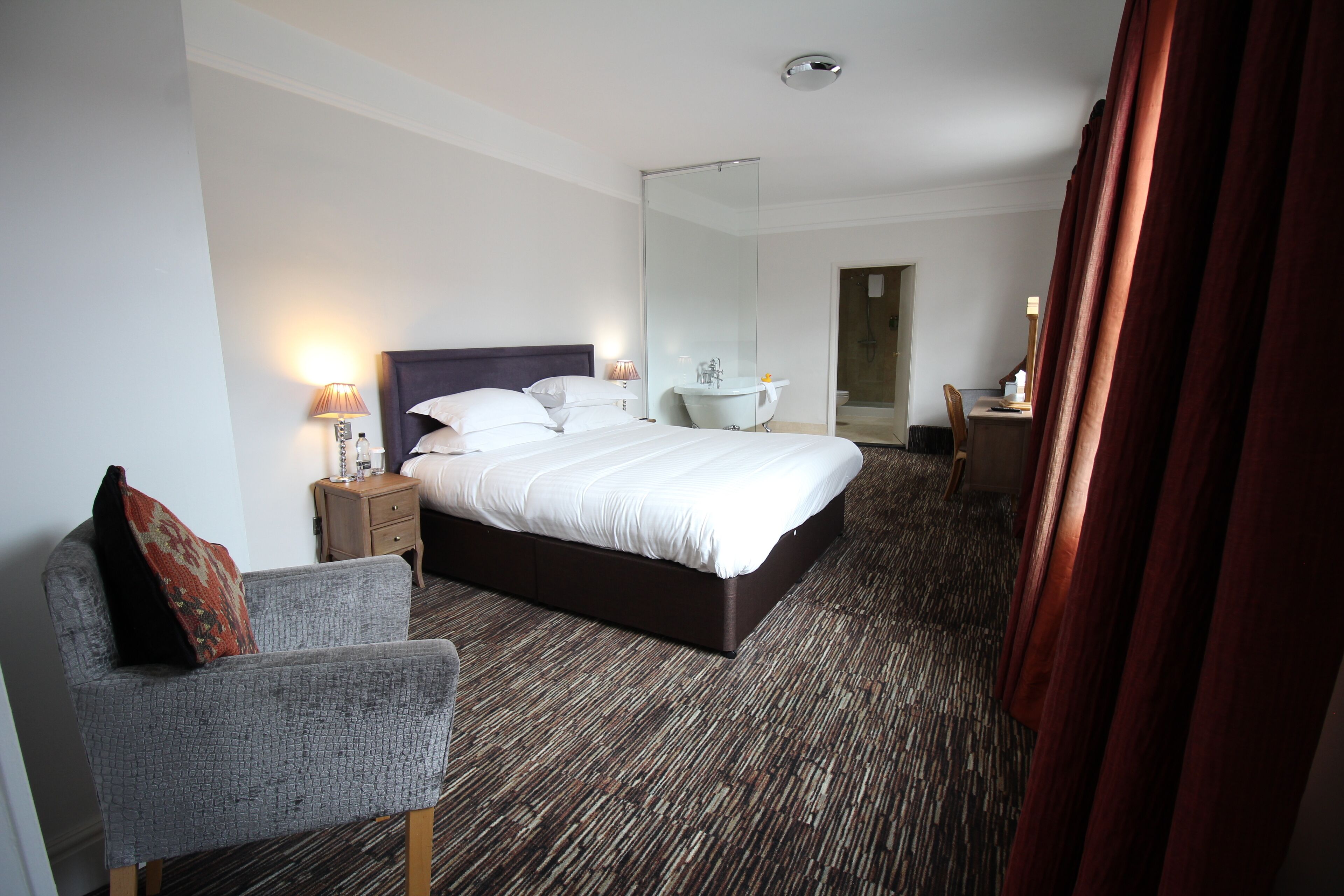 A modern hotel room with a large bed, a sitting area with a chair, a desk, a bathroom with a sink and mirror, and a shower area.