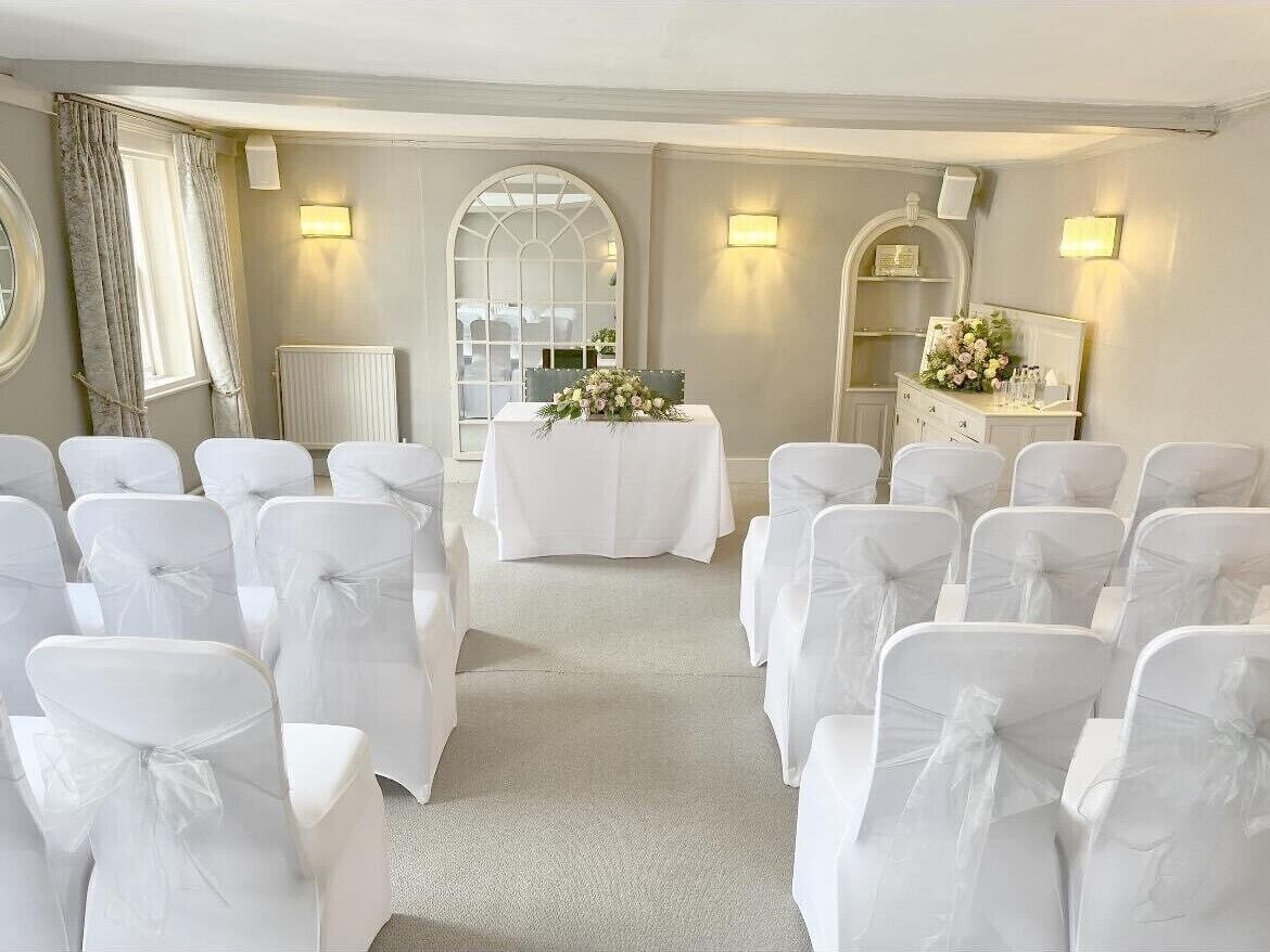 Ein Raum mit Reihen weißer Stühle mit Schleifen, einem Tisch mit Blumenarrangement und einem Spiegelkorpus.