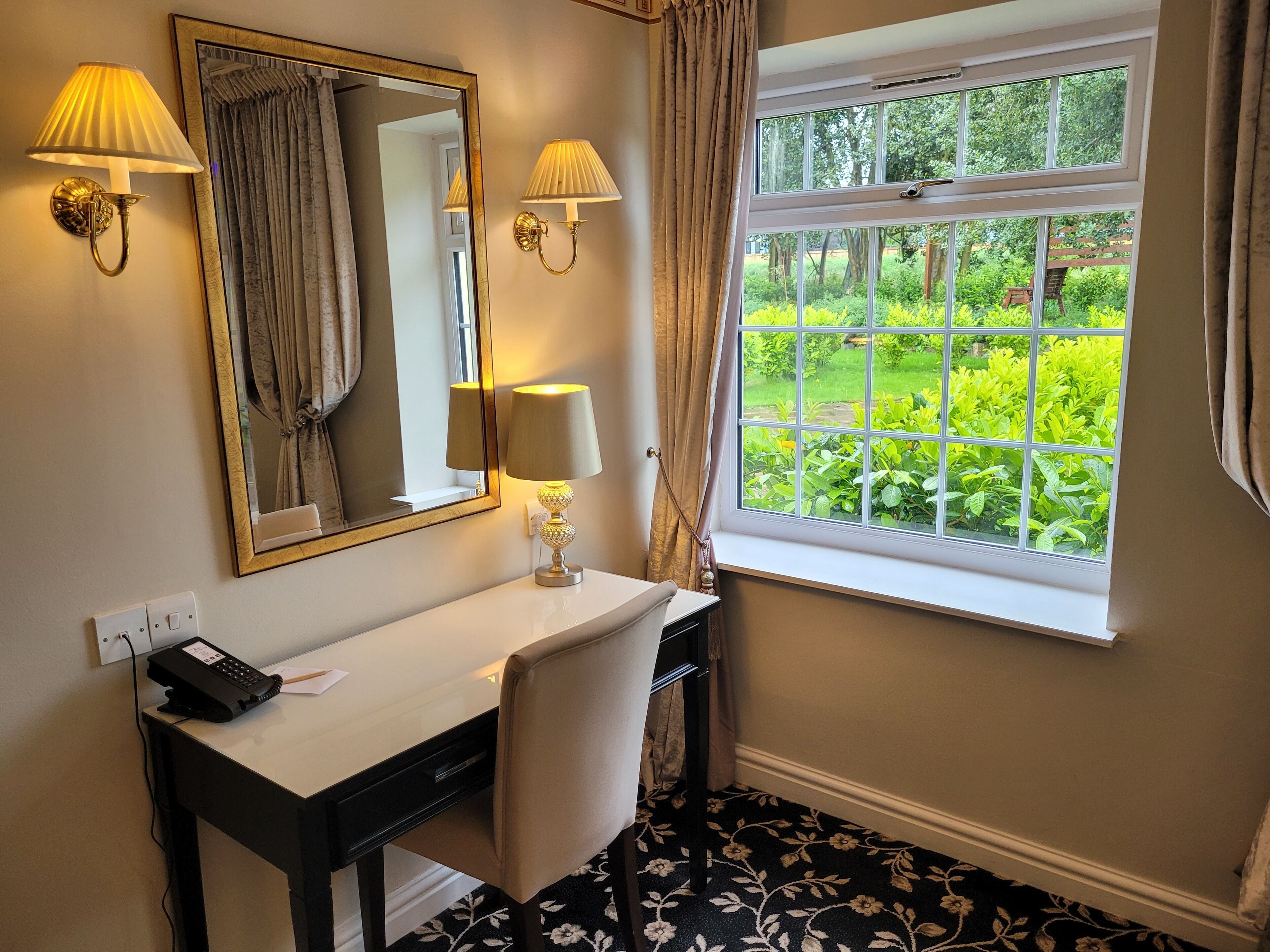 A hotel bathroom with a white vanity, two wall-mounted lamps, a large mirror, and a window with a view of greenery.