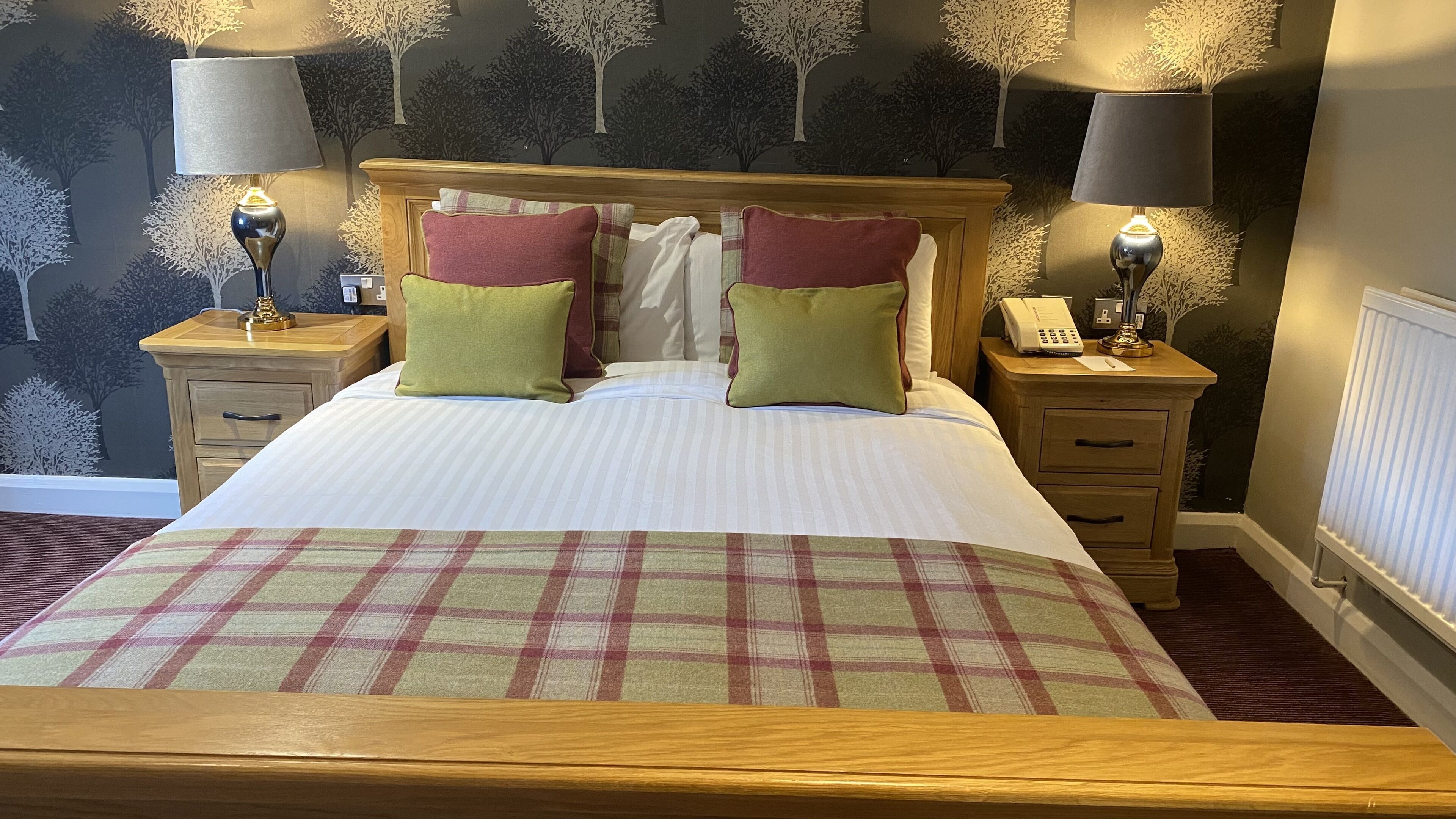 A neatly made bed with a plaid bedspread, two pillows, and a wooden headboard. There are two bedside tables with lamps and a telephone on one of them. The room has a dark wall with a tree mural.