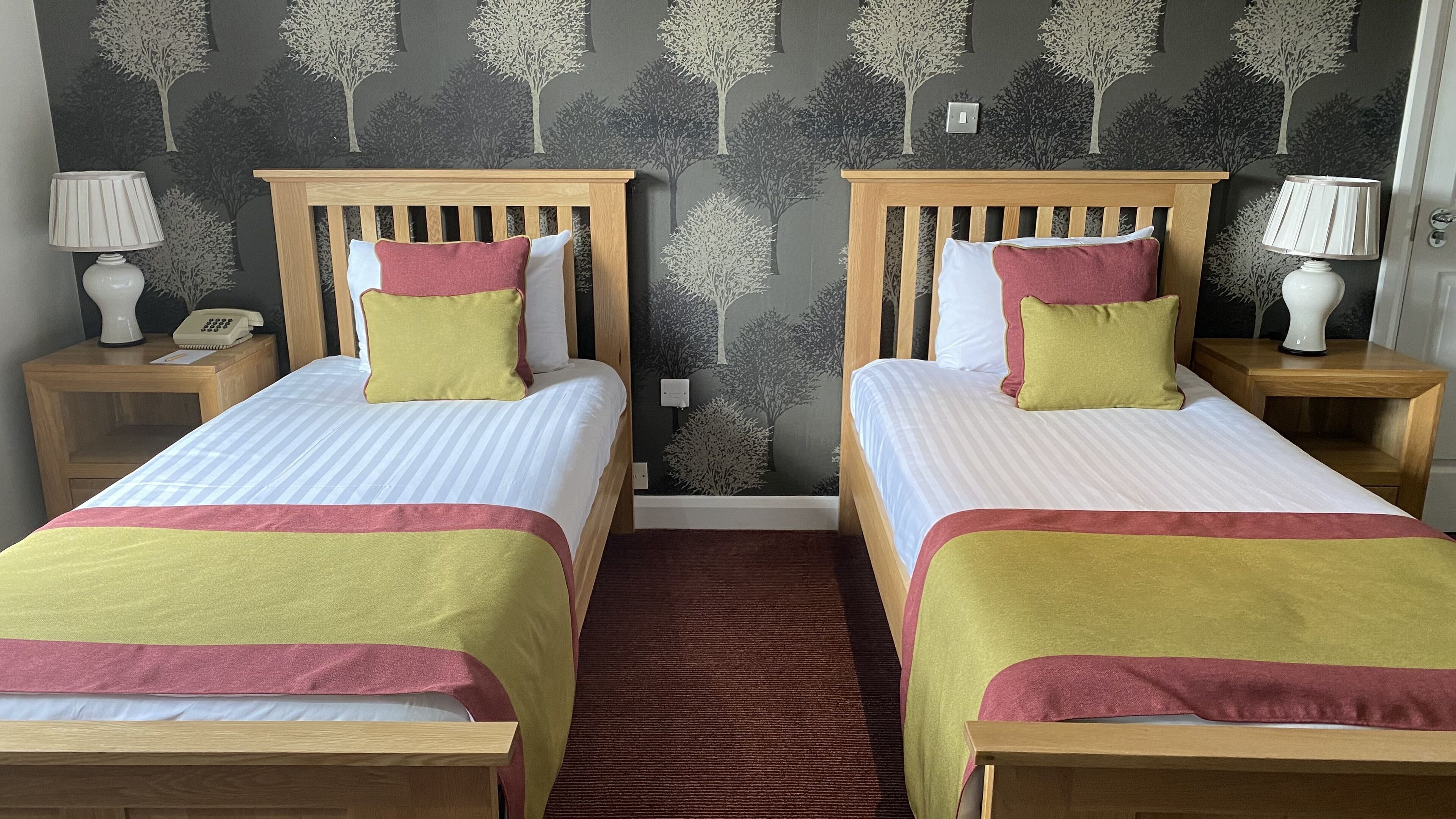 A double room with two beds, each with a wooden headboard, white bedding, and a red and yellow throw. There are bedside tables with lamps and a telephone on one side.