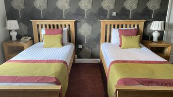 A double room with two beds, each with a wooden headboard, white bedding, and a red and yellow throw. There are bedside tables with lamps and a telephone on one side.