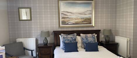 A hotel room with a bed, two bedside tables, a chair, a radiator, and a painting on the wall.