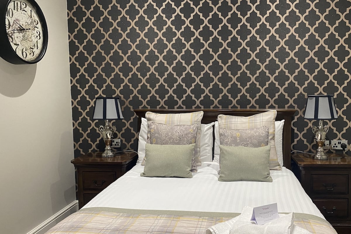 Quarto de hotel com papel de parede estampado, cama com roupa de cama branca, duas mesas de cabeceira com abajures e um relógio na parede.