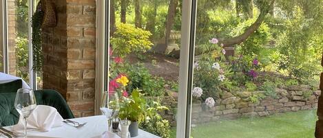 A dining area with a table set for two, a blue chair, and a view of a garden through large windows.