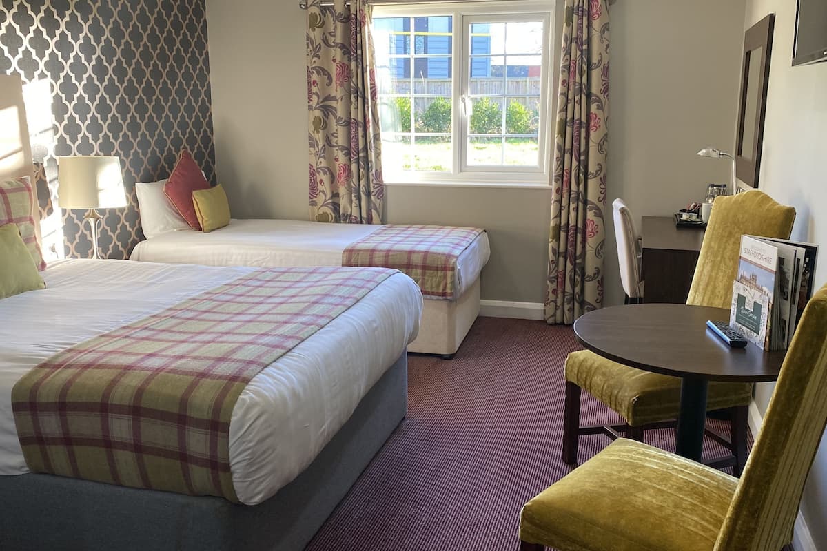 Quarto de hotel com duas camas, uma mesa redonda pequena, uma cadeira, uma janela com cortinas e uma televisão na parede.