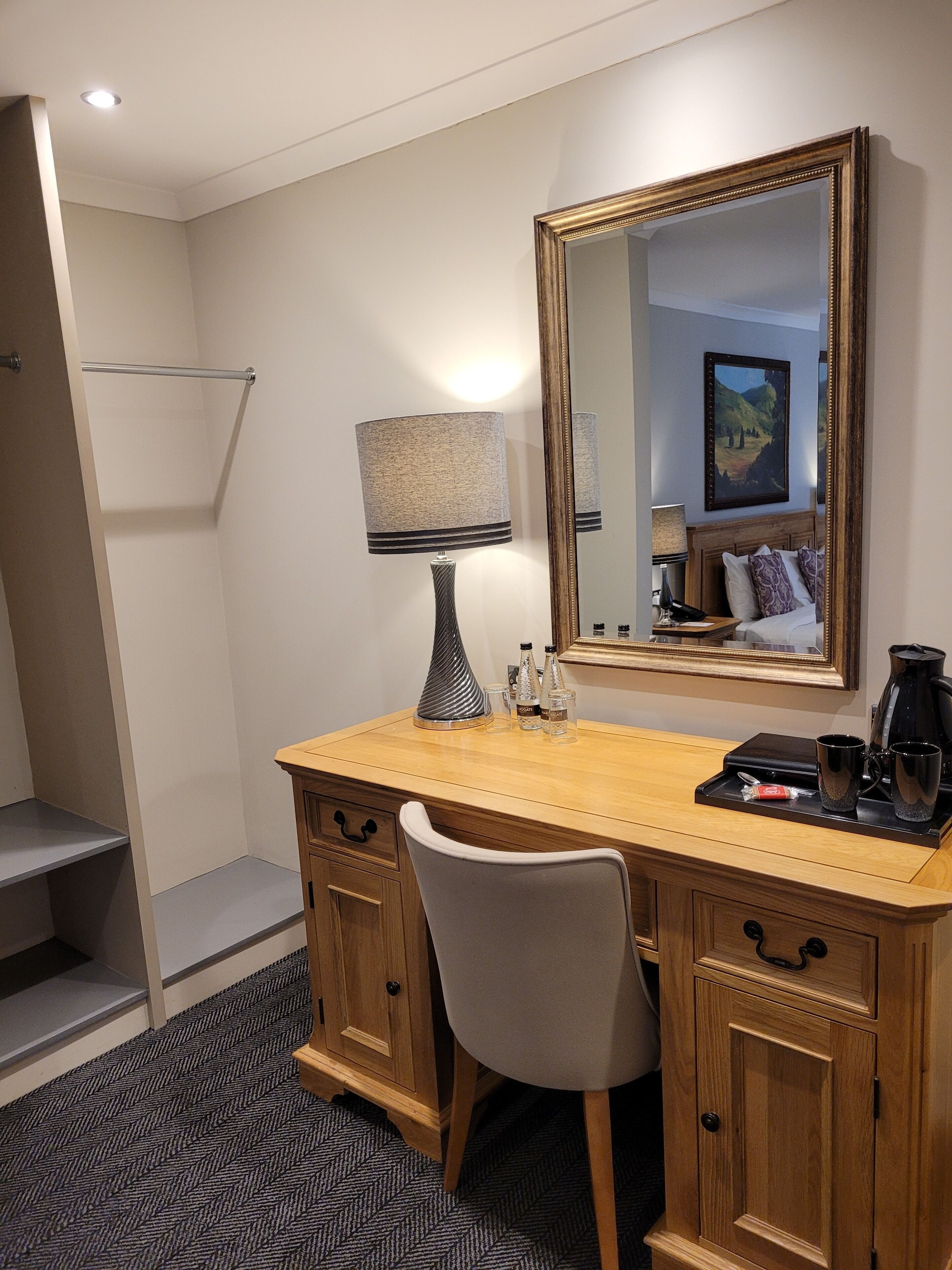 A hotel room with a wooden desk, a chair, a large mirror, a lamp, a bed with pillows, a coffee maker, and a small table with two cups and a bottle.