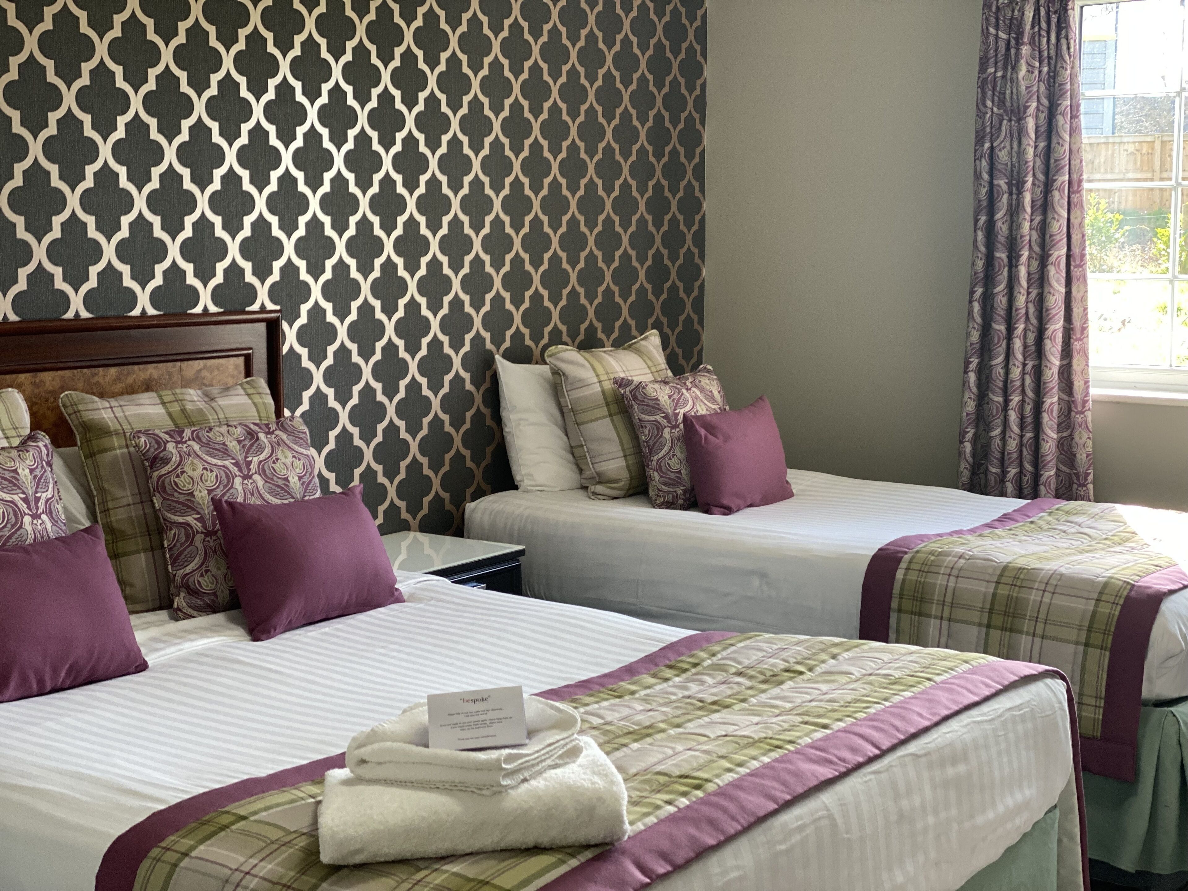 A hotel room with two beds, each with a headboard, multiple pillows, a folded towel, and a note. The room has patterned wallpaper and a window with curtains.