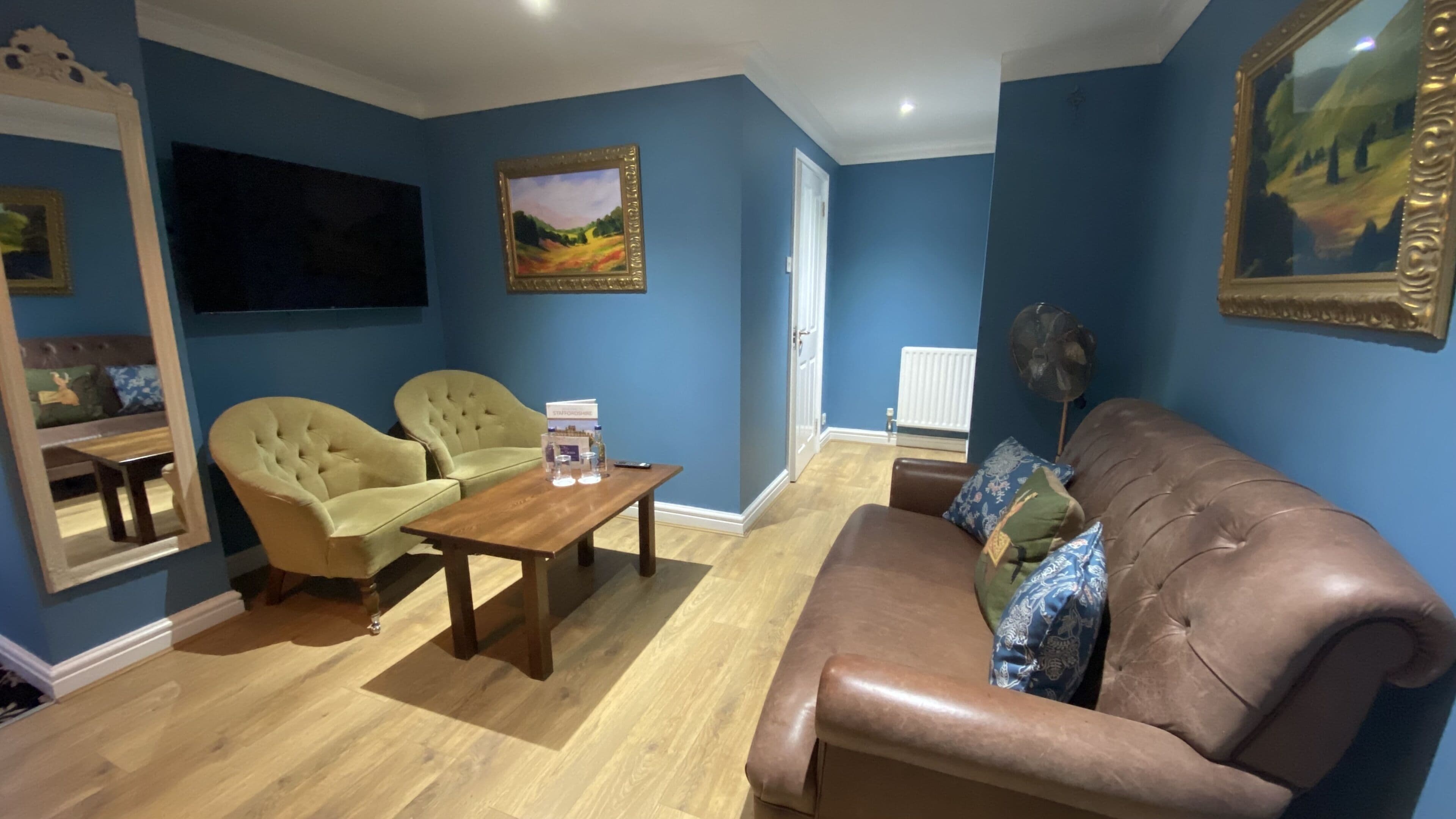 Ein Wohnzimmer mit blauen Wänden, einem braunen Ledersofa, zwei grünen Sesseln, einem Holztisch und einem großen Spiegel.