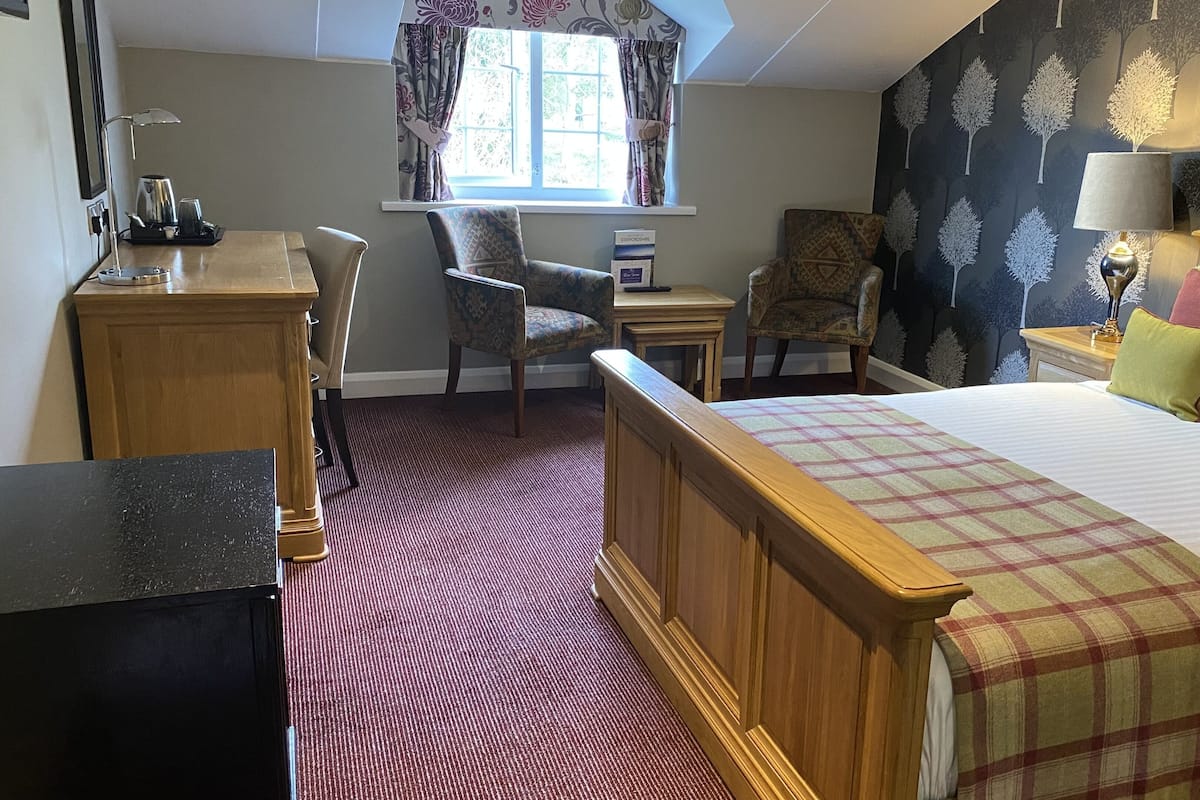 Quarto de hotel com cama de madeira, uma escrivaninha com cadeira, uma mesinha e janela com cortinas.