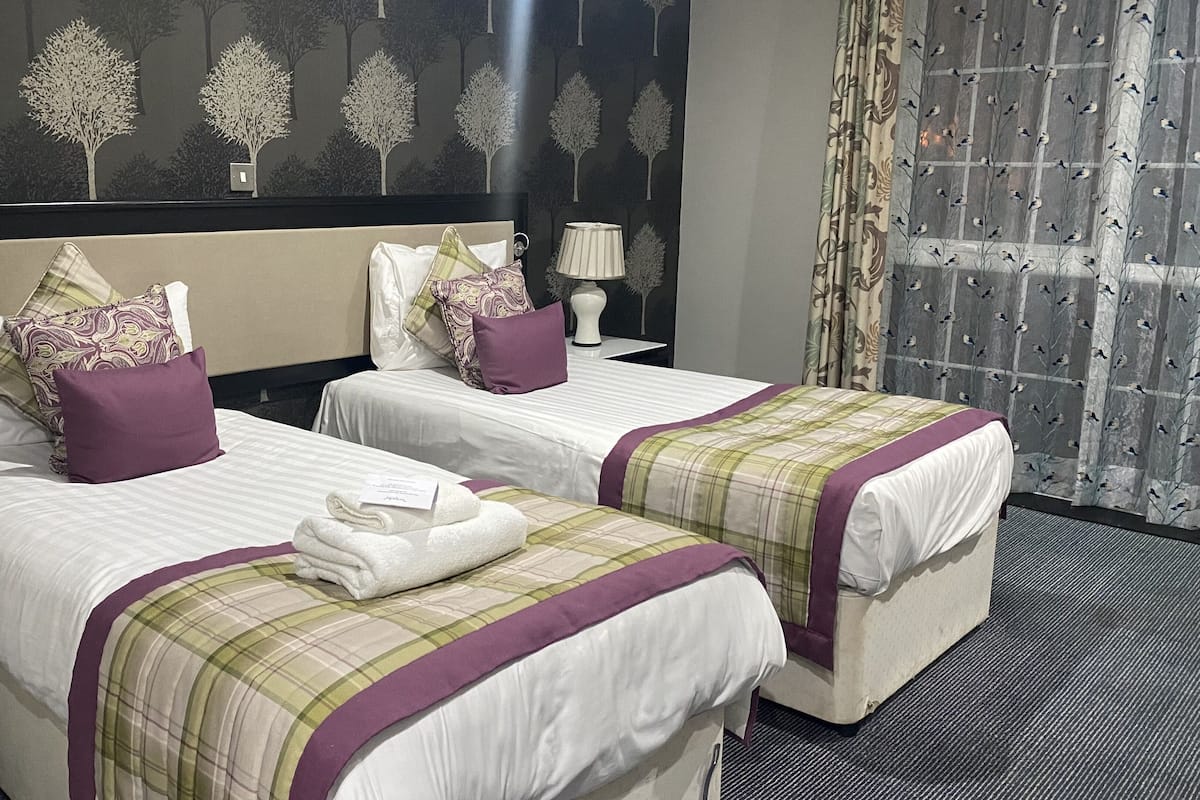 Quarto de hotel com duas camas, cada uma com cabeceira, almofadas e manta. Há uma luminária em uma mesinha de cabeceira e um telefone no criado-mudo.